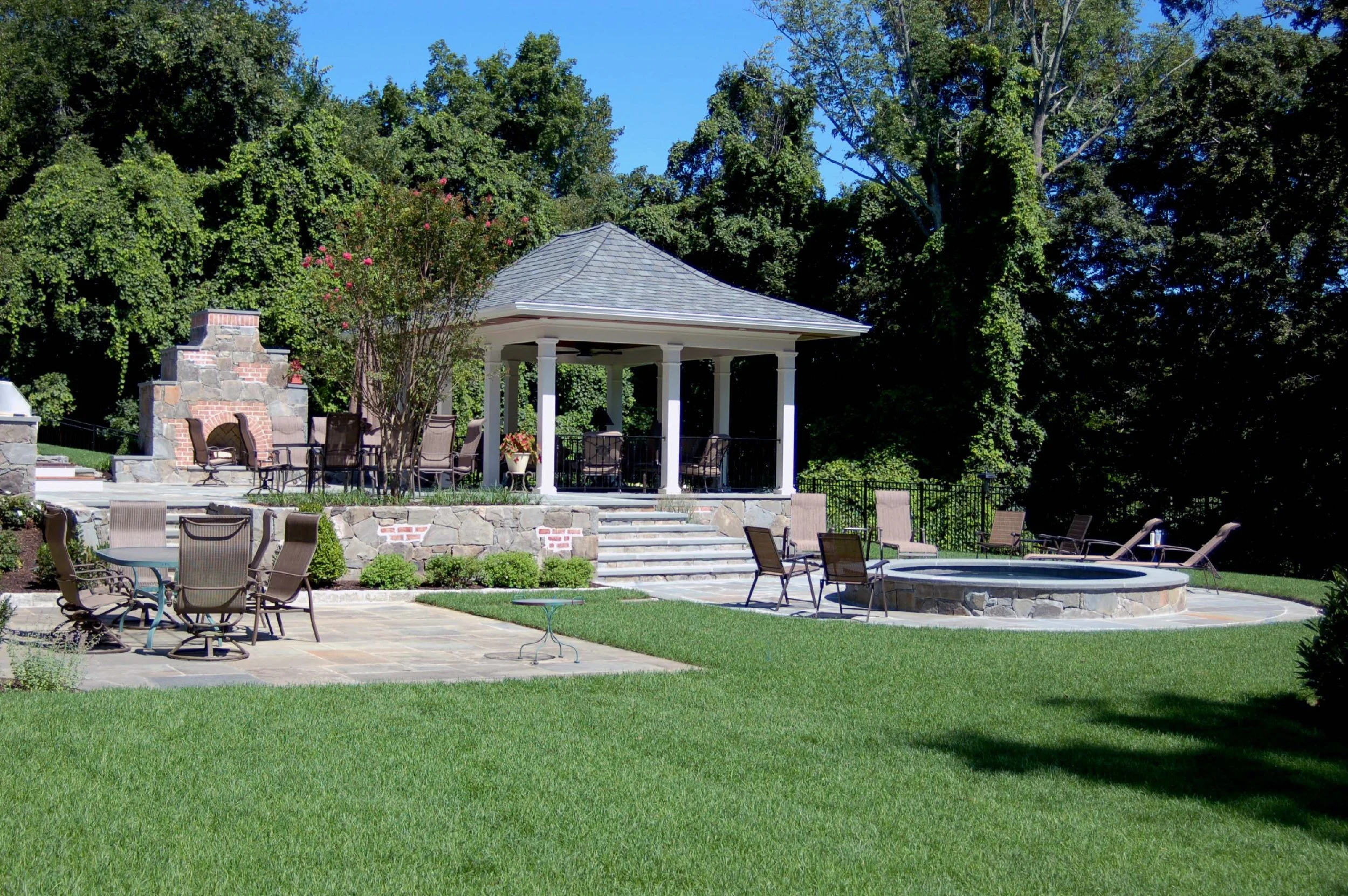 Backyard patio with stone fireplace, a gazebo with chairs, and a circular hot tub, surrounded by green grass and trees on a sunny day.