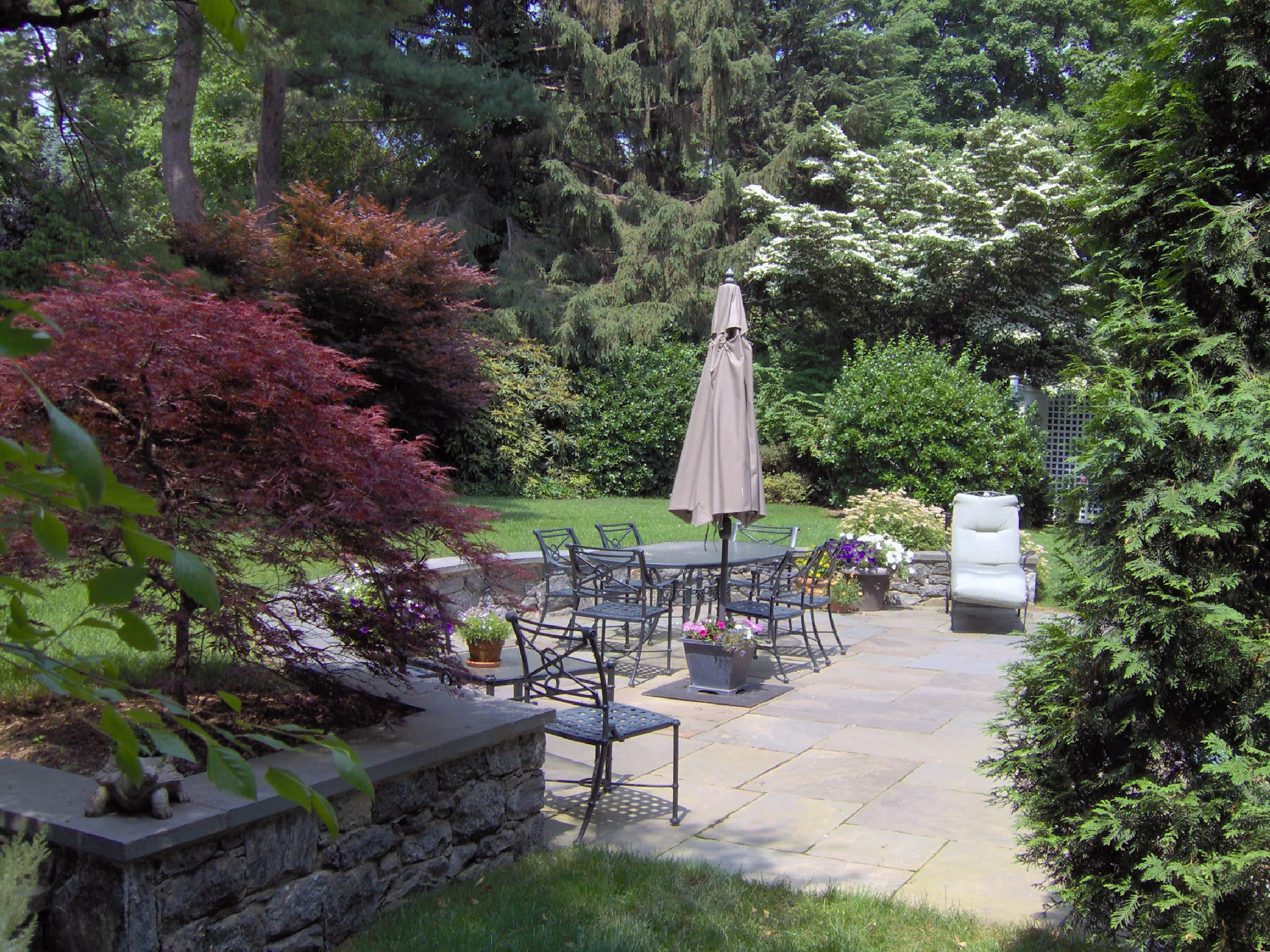 A lush backyard patio with stone pavers, surrounded by green trees and shrubs. There is a round metal table with chairs, an umbrella, potted flowers, and a white reclining chair.