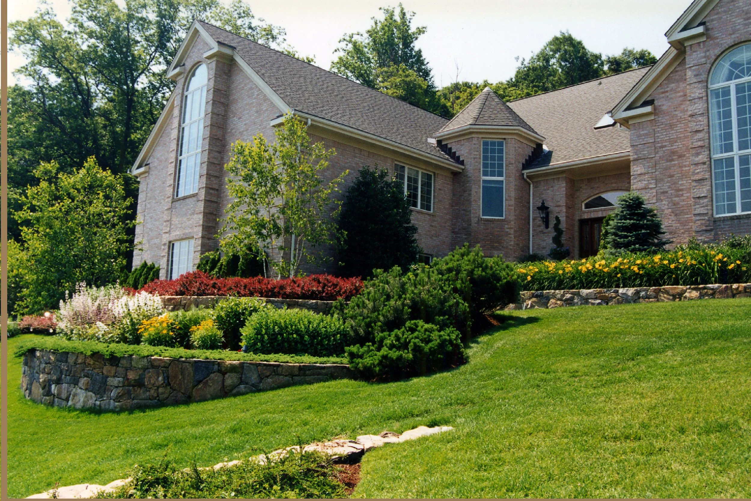 A large brick house with multiple windows surrounded by a landscaped garden with colorful flowers and green shrubs, on a sunny day with trees in the background.
