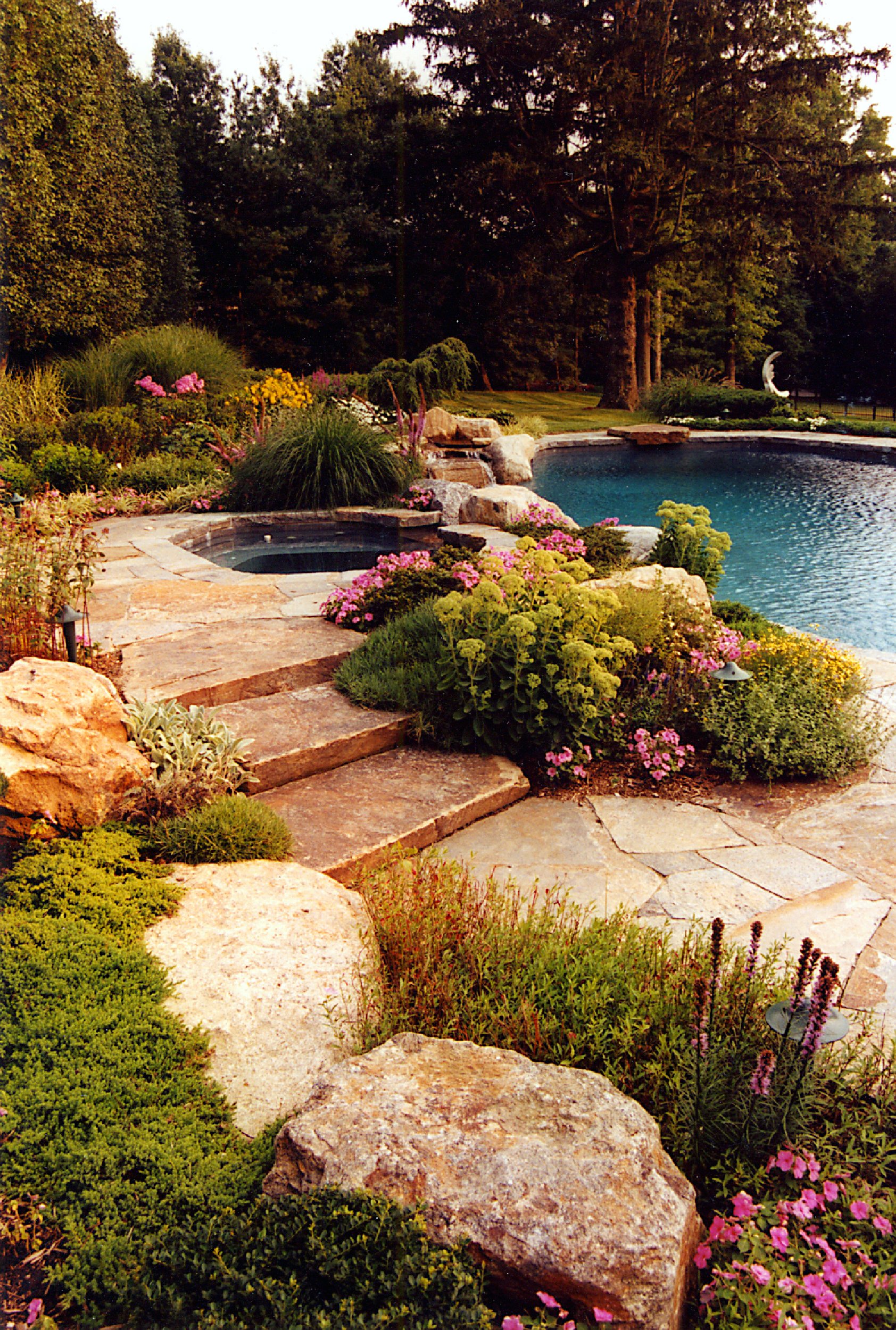 A backyard pool area with a stone patio, surrounded by lush greenery and colorful flowers, with trees and a sculpture in the background.
