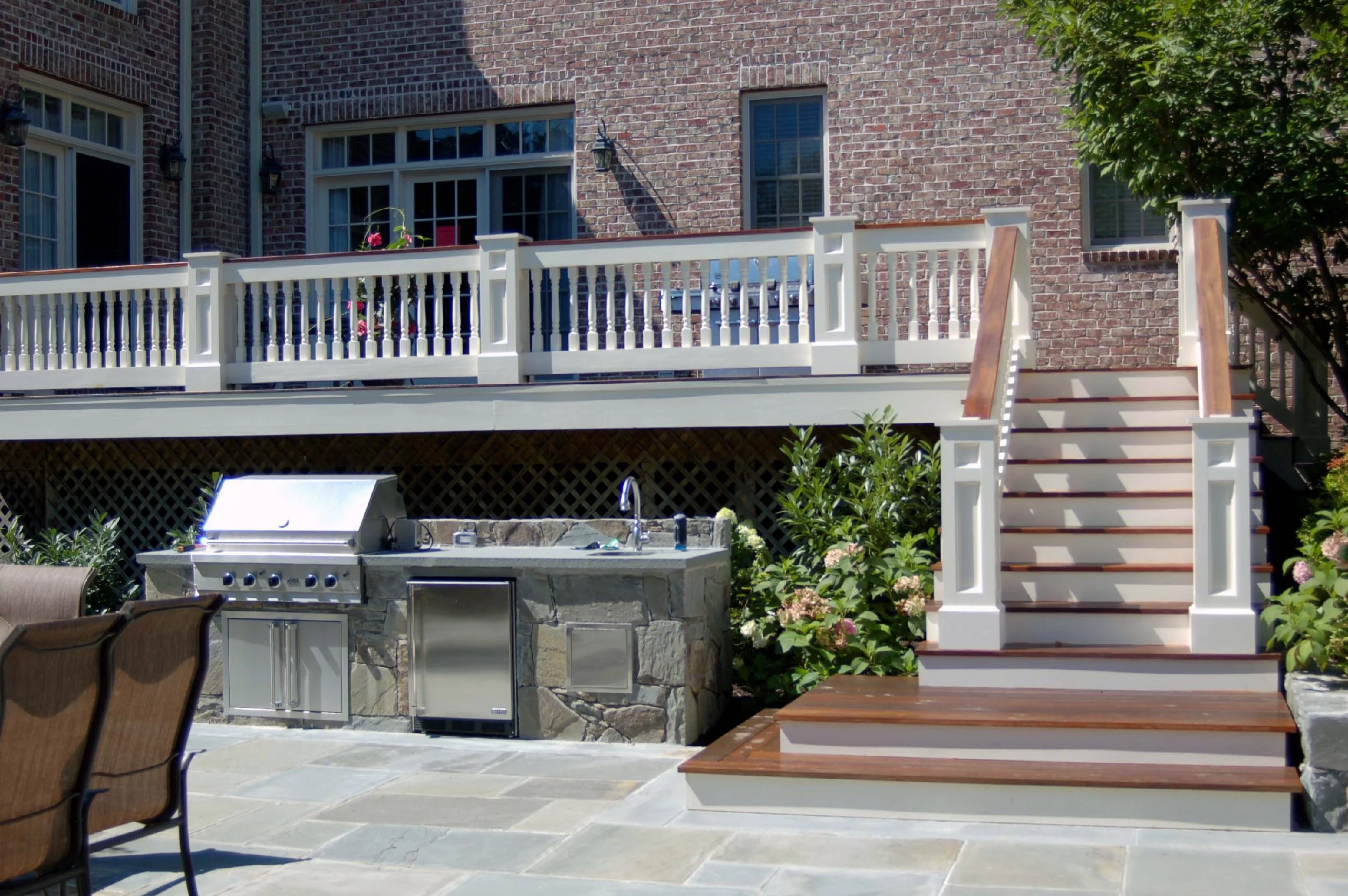 Backyard with a deck and staircase, outdoor kitchen with a grill, and patio seating area with chairs, surrounded by landscaping and flowering plants.