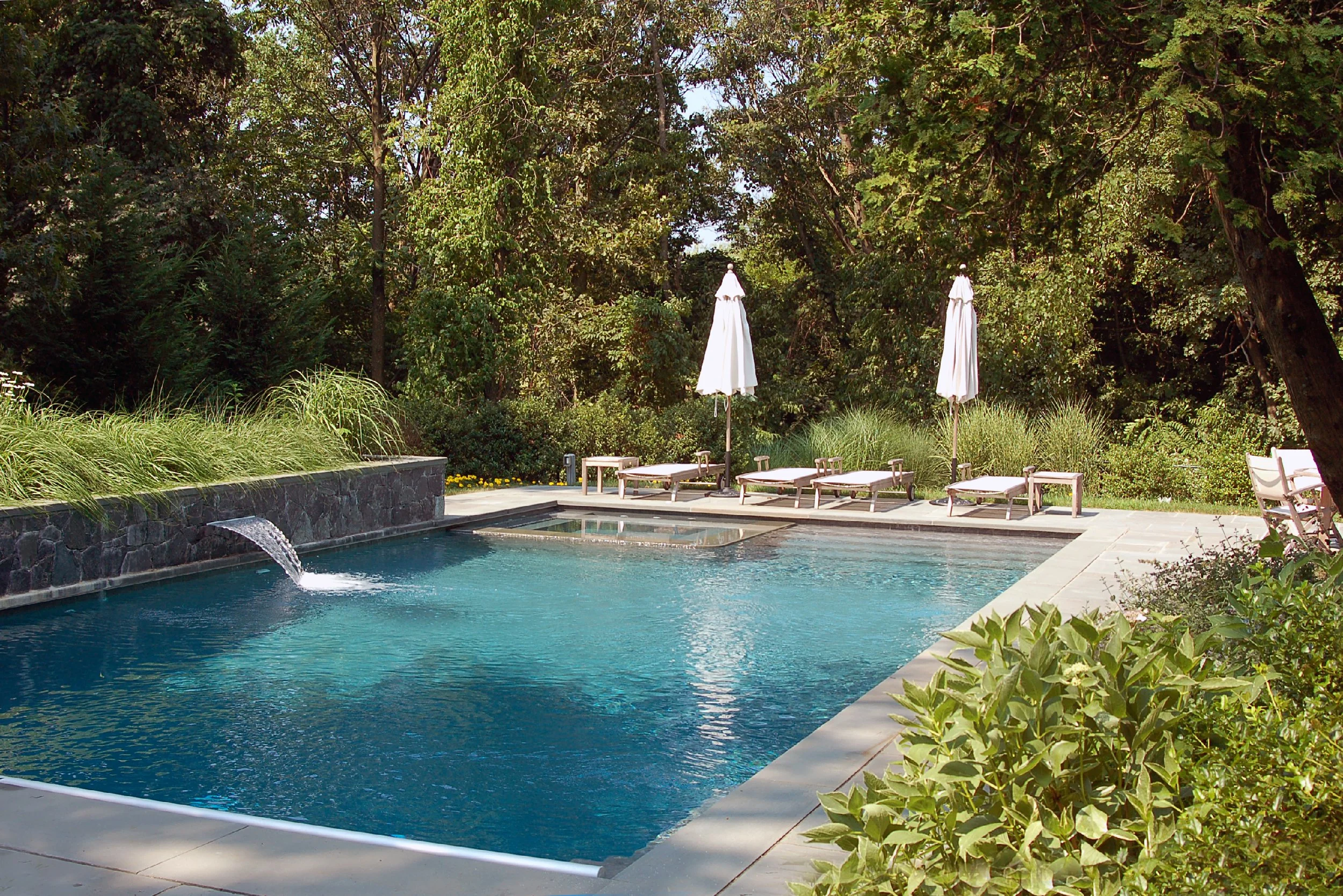 A backyard swimming pool surrounded by greenery, with a stone waterfall feature flowing into the pool, poolside chairs, and white umbrellas under trees.