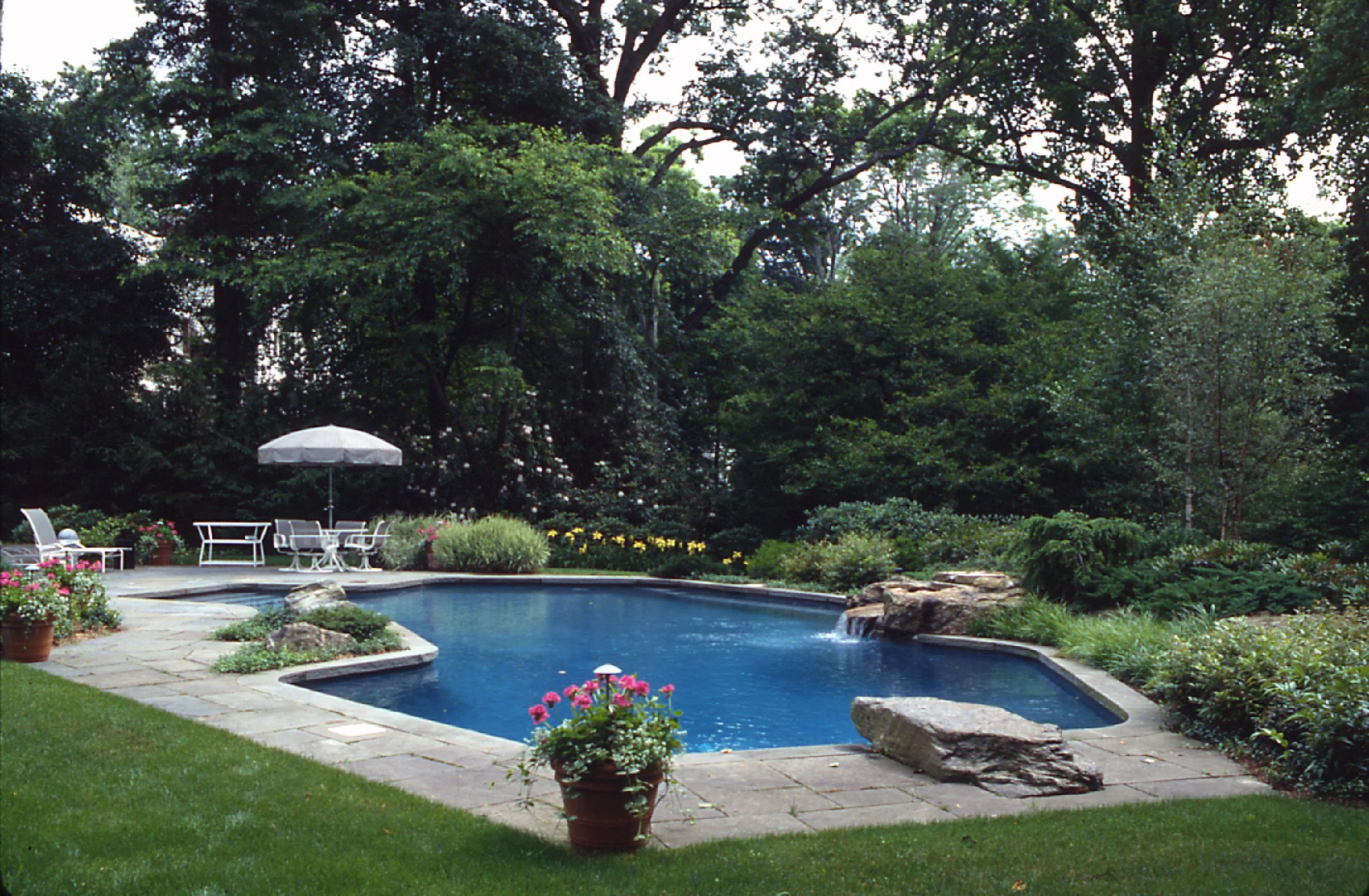 Backyard with a rectangular swimming pool, surrounded by paving stones, potted plants with pink flowers, garden beds with bushes and trees, and outdoor furniture including a white table with an umbrella and chairs.