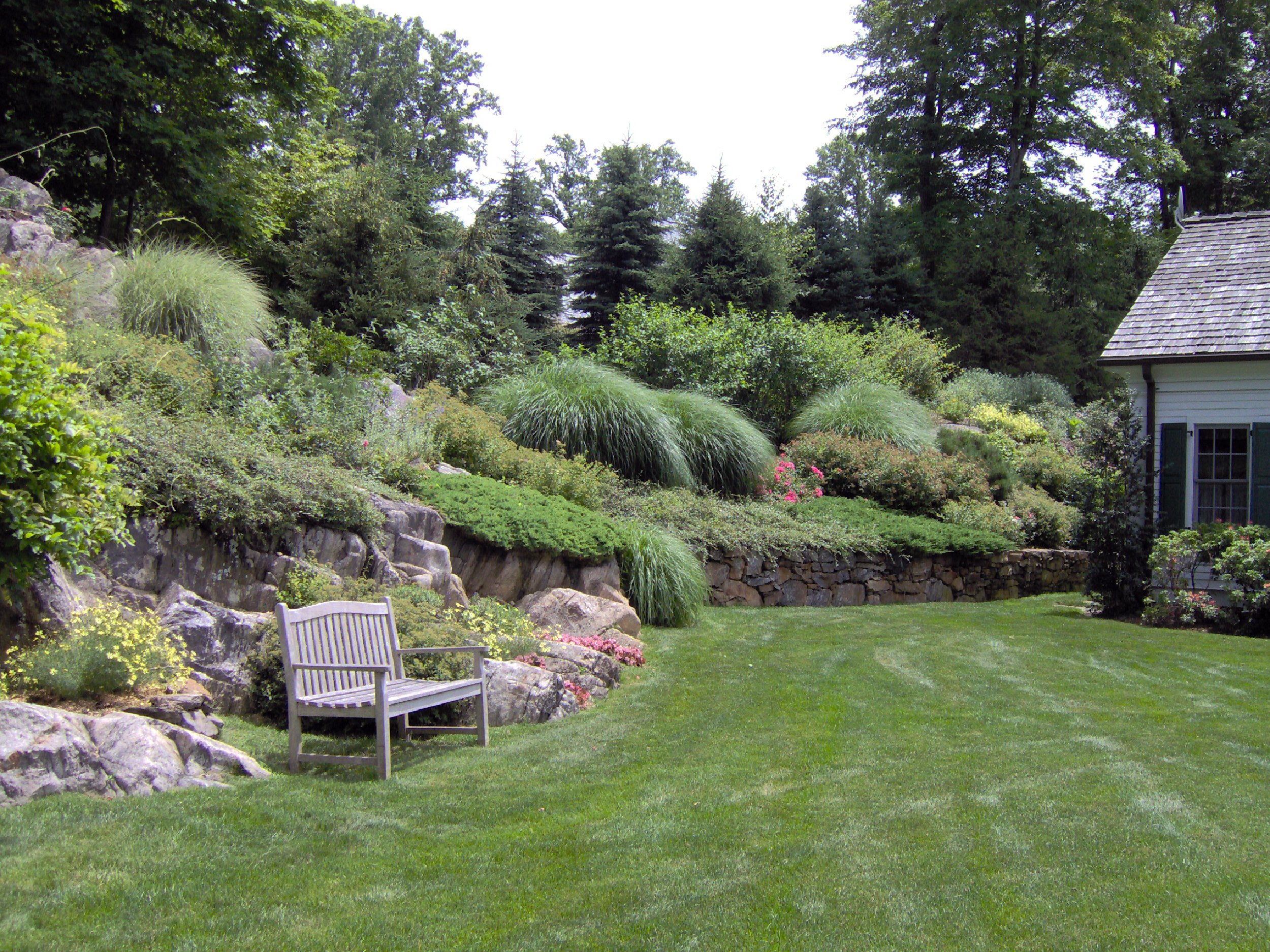 Lush backyard garden with well-maintained green lawn, various shrubs, and tall trees in the background. A wooden bench is positioned on the grass near a rocky, landscaped slope with different plants and flowers. A house with white siding and dark shutters is partially visible on the right.