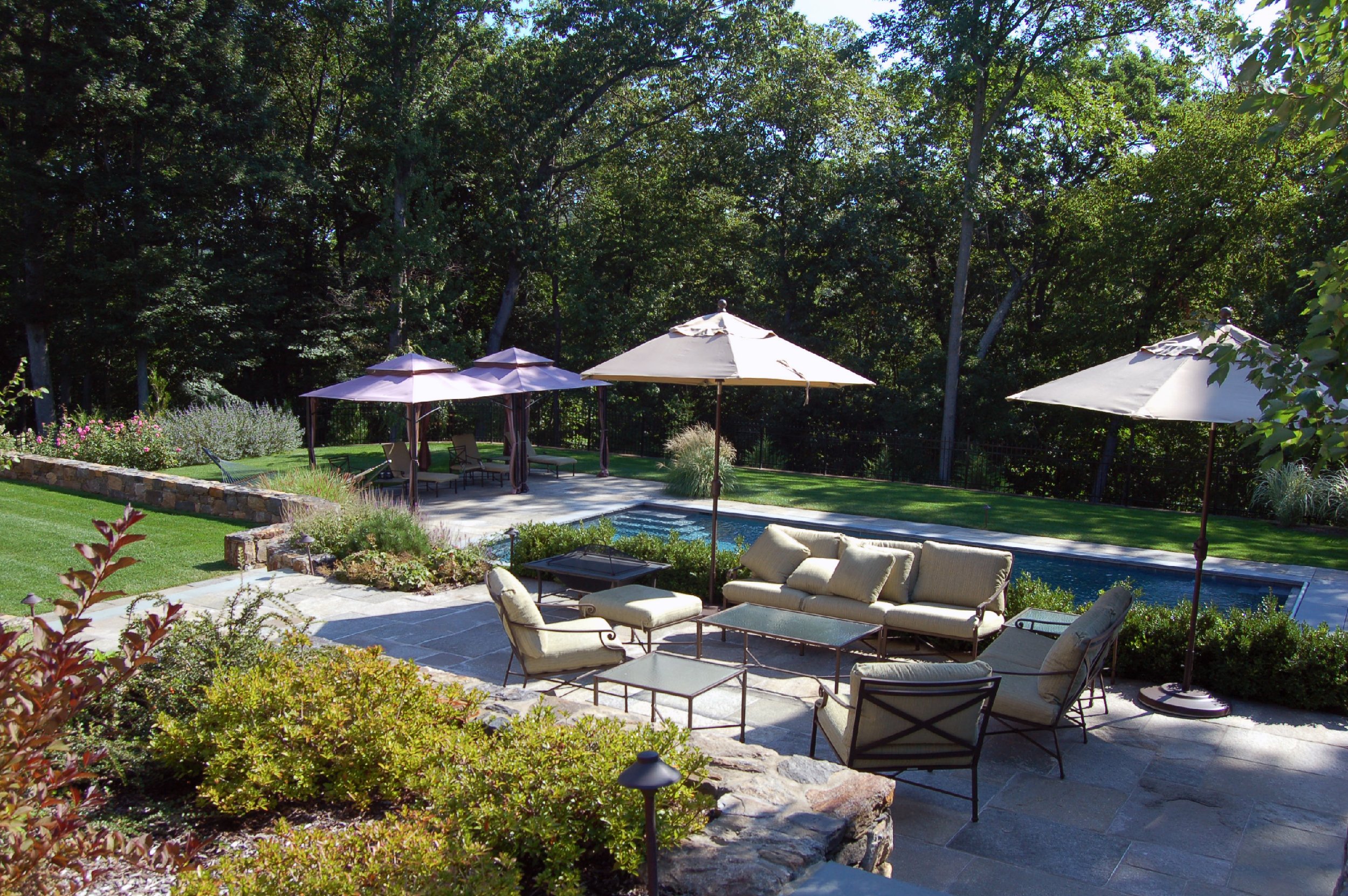 Outdoor patio area with beige cushioned furniture, glass-top tables, umbrellas, a swimming pool, lush green lawns, flowerbeds, and trees in the background.