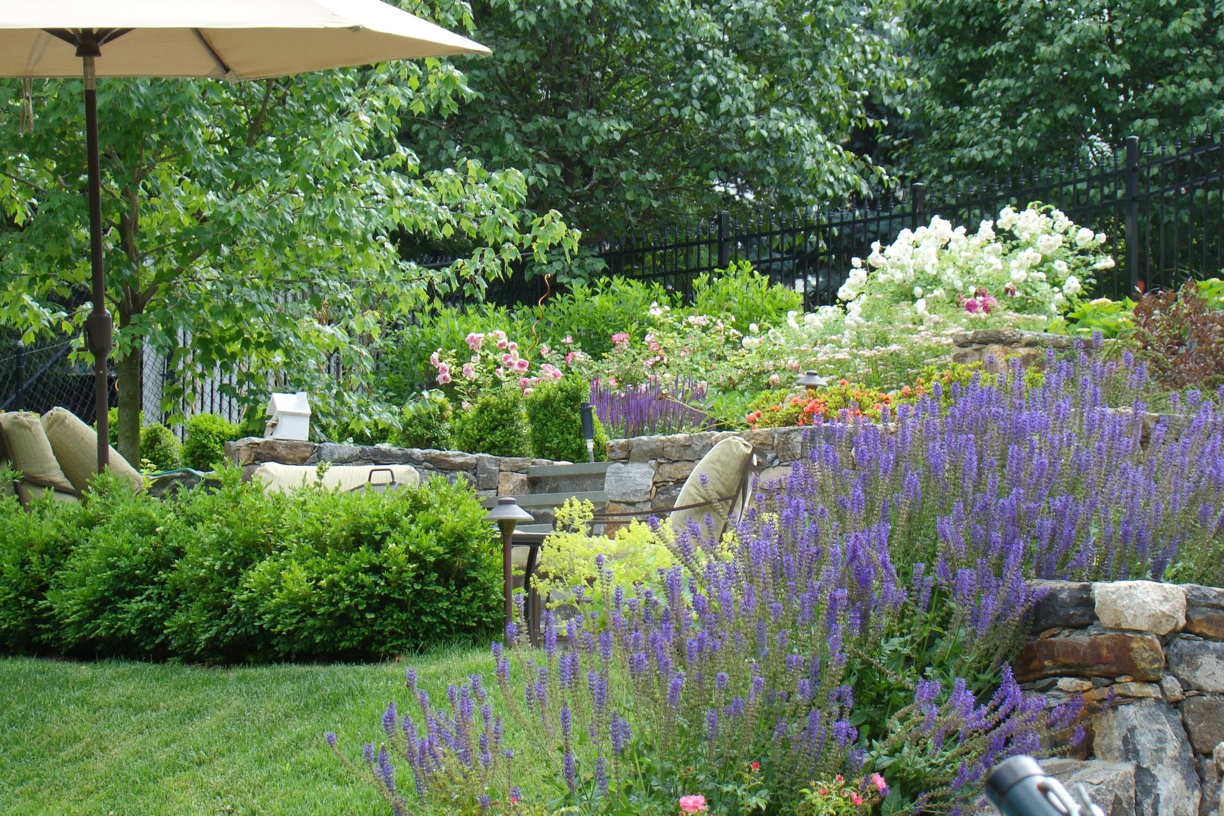 A lush garden with a stone retaining wall filled with colorful flowers including lavender, roses, and other blooming plants, and a patio area with outdoor cushions, an umbrella, and garden lights.