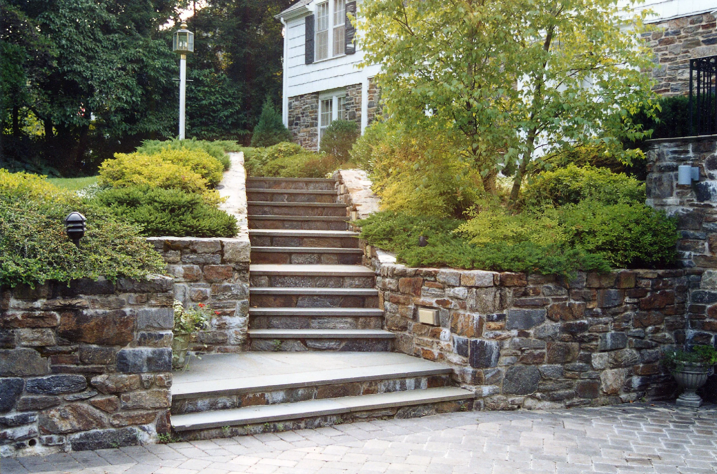 Stone stairs leading to a house with siding and window shutters, surrounded by green shrubs and trees, with pathway and garden lighting.