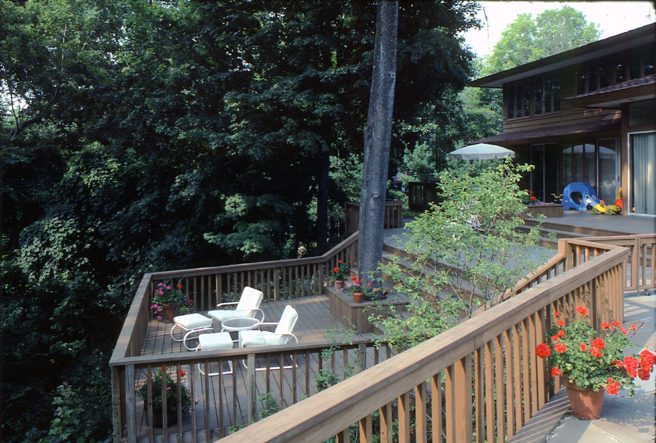 Backyard patio with wooden railing, seating area with white chairs, potted plants, large trees, and a house with large windows and children's toys on the deck.