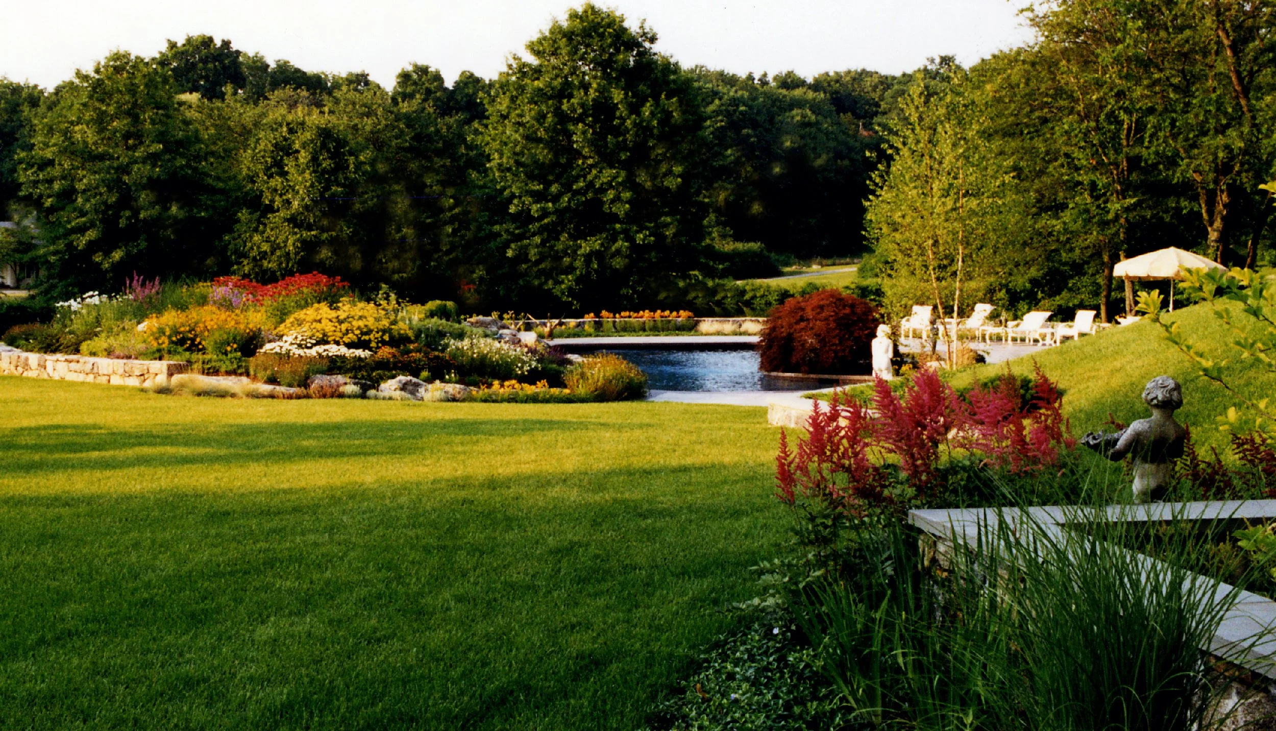 A landscaped backyard with a green lawn, colorful flower beds, a pond, and outdoor seating area with white chairs and umbrella, surrounded by trees and decorative statues.