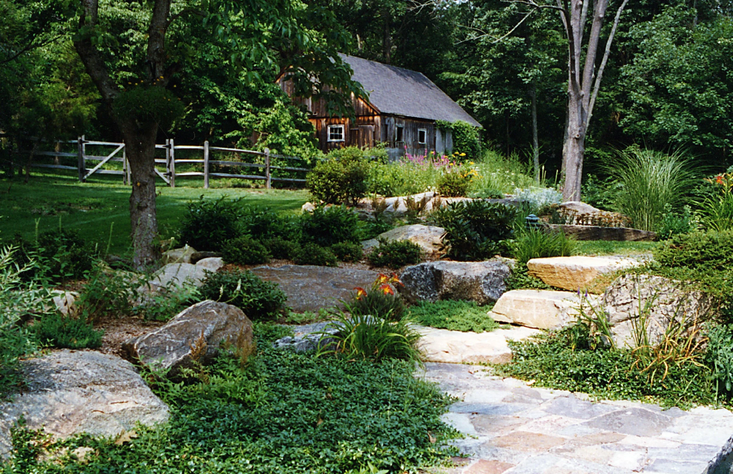 A lush garden with large rocks, green plants, and flowers, with a rustic wooden shed in a wooded area.