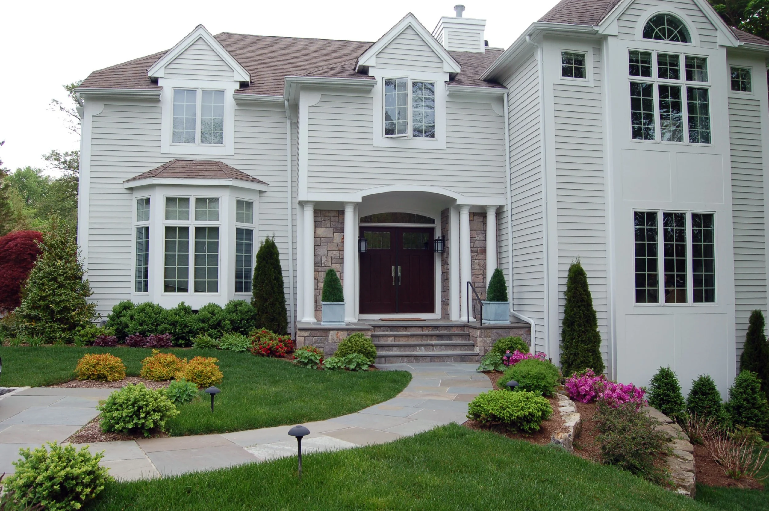 Front view of a large, white, two-story house with multiple windows, a brown door, and a landscaped lawn with bushes, flowers, and pathway.