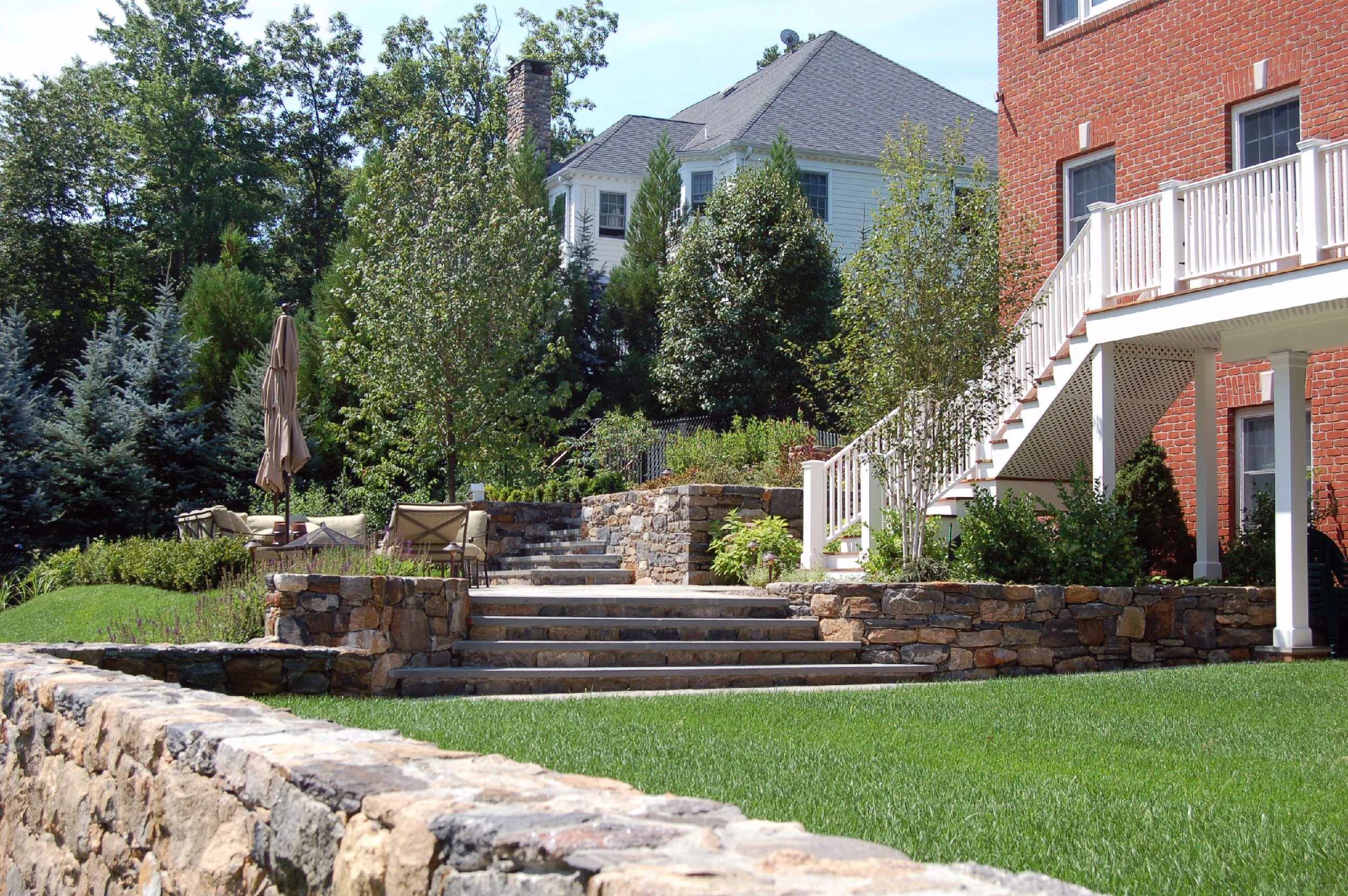 Backyard with stone stairs, patio furniture, lush green lawn, and garden surrounded by trees and shrubs, brick and white house in the background.