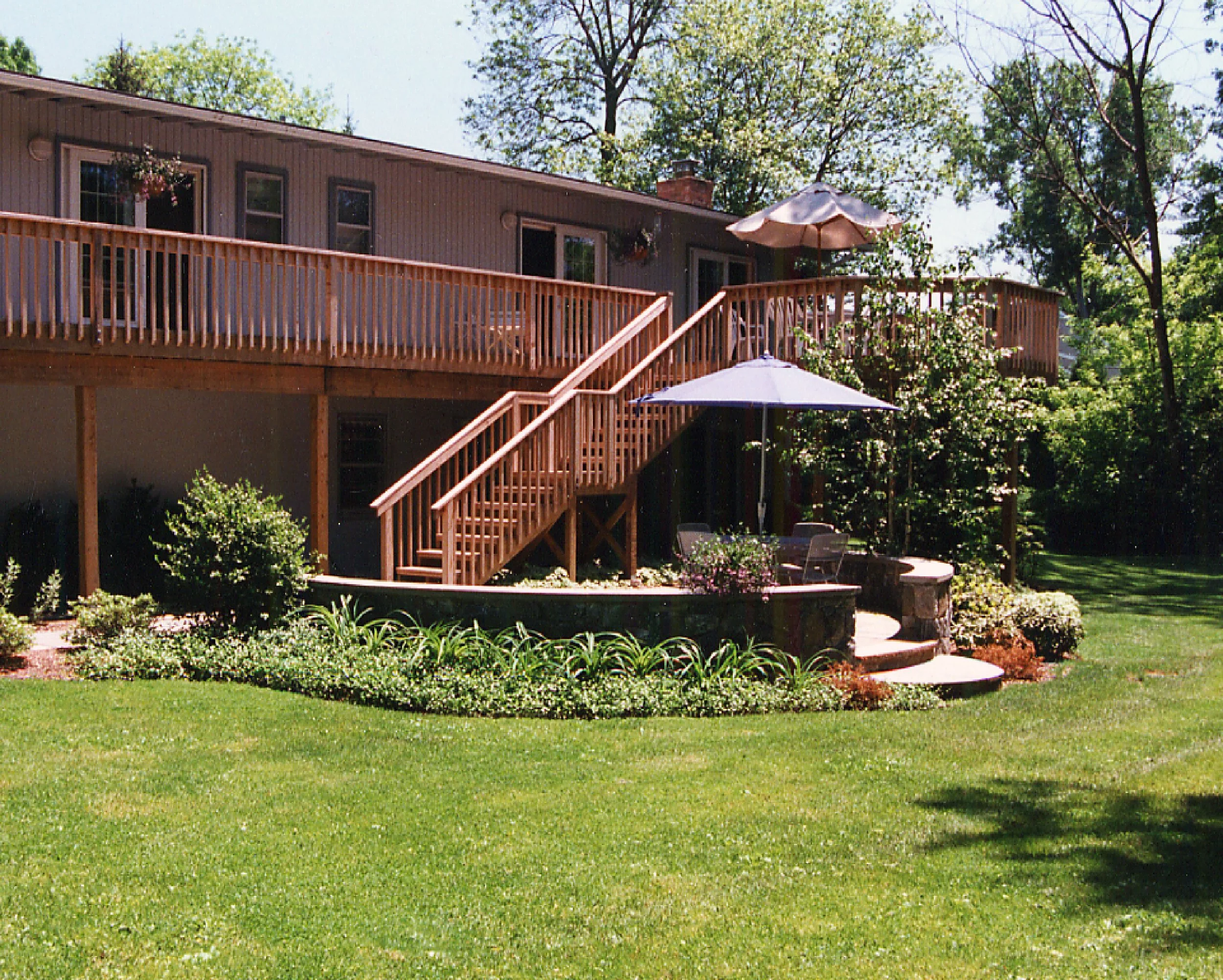 Backyard with a wooden deck, staircase, patio furniture, umbrellas, lush landscaping, and trees in the background.
