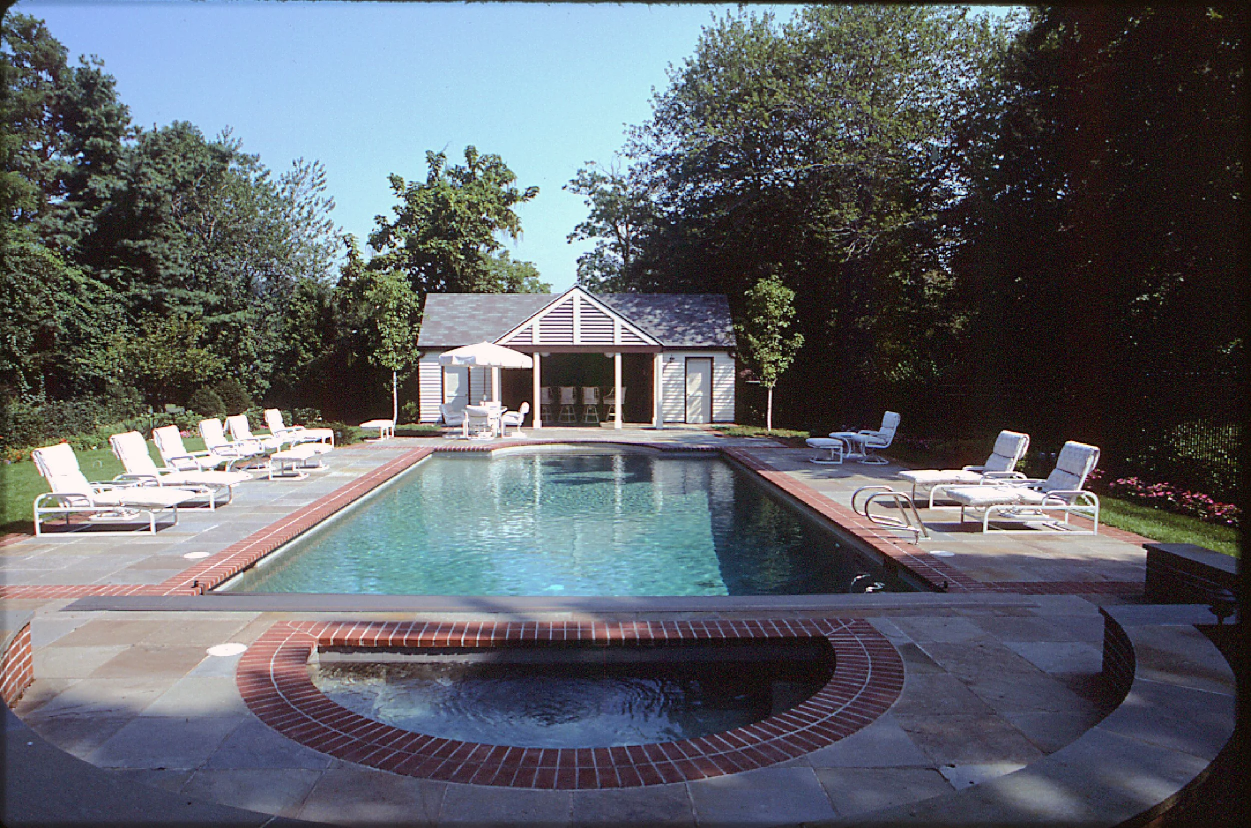 A backyard swimming pool surrounded by lounge chairs, with a small pool house or cabana at the far end, set amidst trees and greenery on a sunny day.