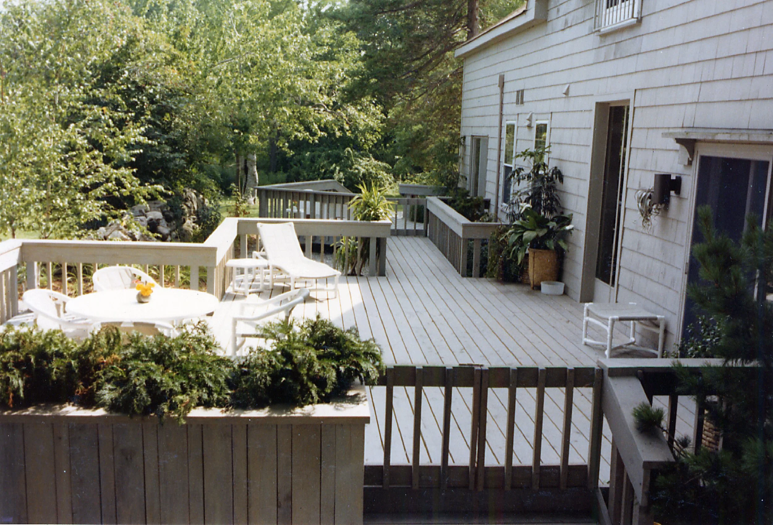Wooden outdoor deck attached to a white house with sliding glass door, surrounded by houseplants and outdoor furniture, with a lush green backyard in the background.