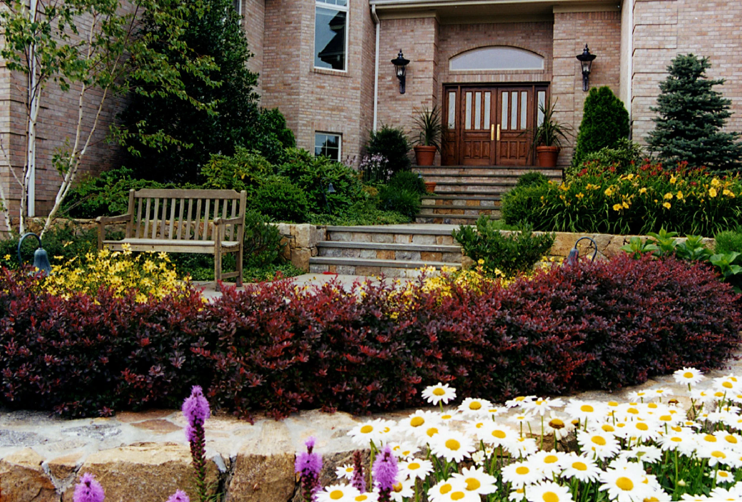 A landscaped front yard with a stone pathway and steps leading up to a brick house with wooden double doors. The yard features various bushes, flowers, and a wooden bench.