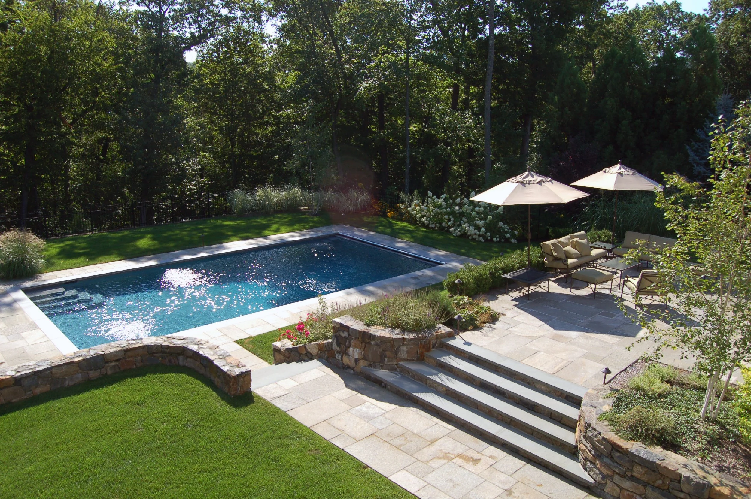 A backyard with a rectangular swimming pool, stone steps, green lawn, patio with outdoor furniture and umbrellas, and surrounded by trees and shrubs.