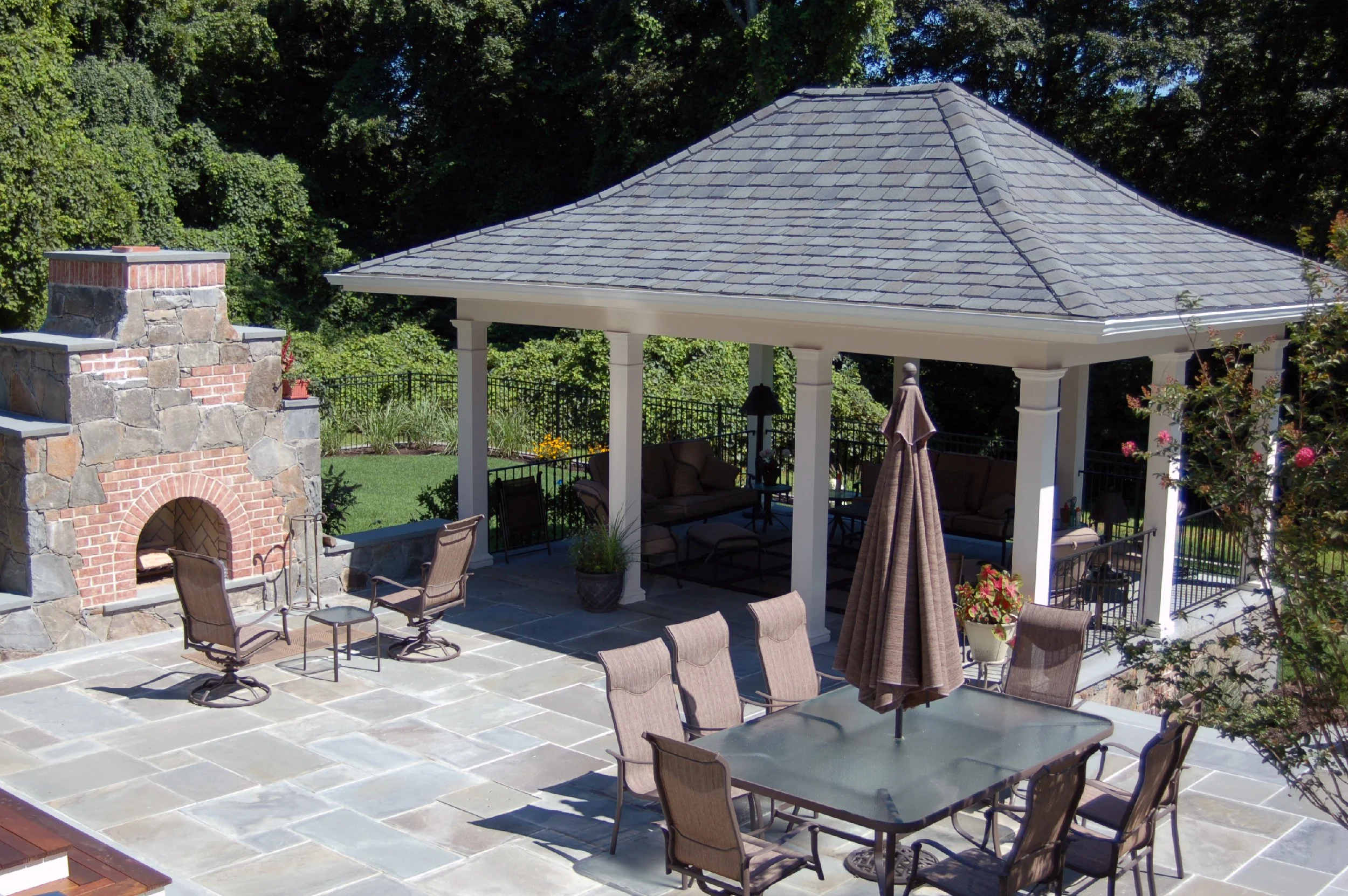 Backyard patio with stone flooring, a stone and brick outdoor fireplace, a dining table with chairs and an umbrella, and a gazebo with outdoor seating and a view of trees.