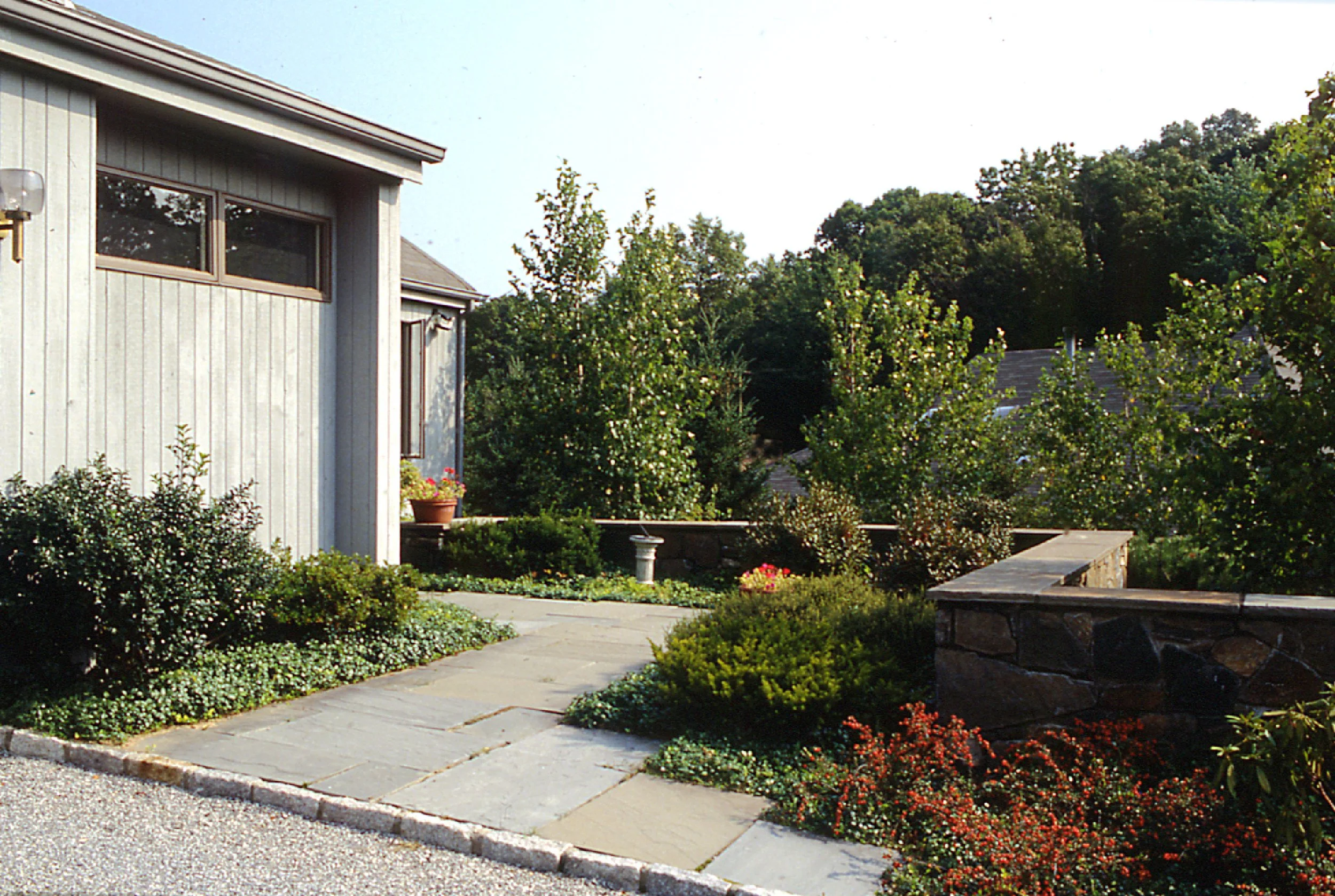A landscaped garden beside a house with a stone pathway, green shrubs, flowering plants, and trees, under a clear sky.