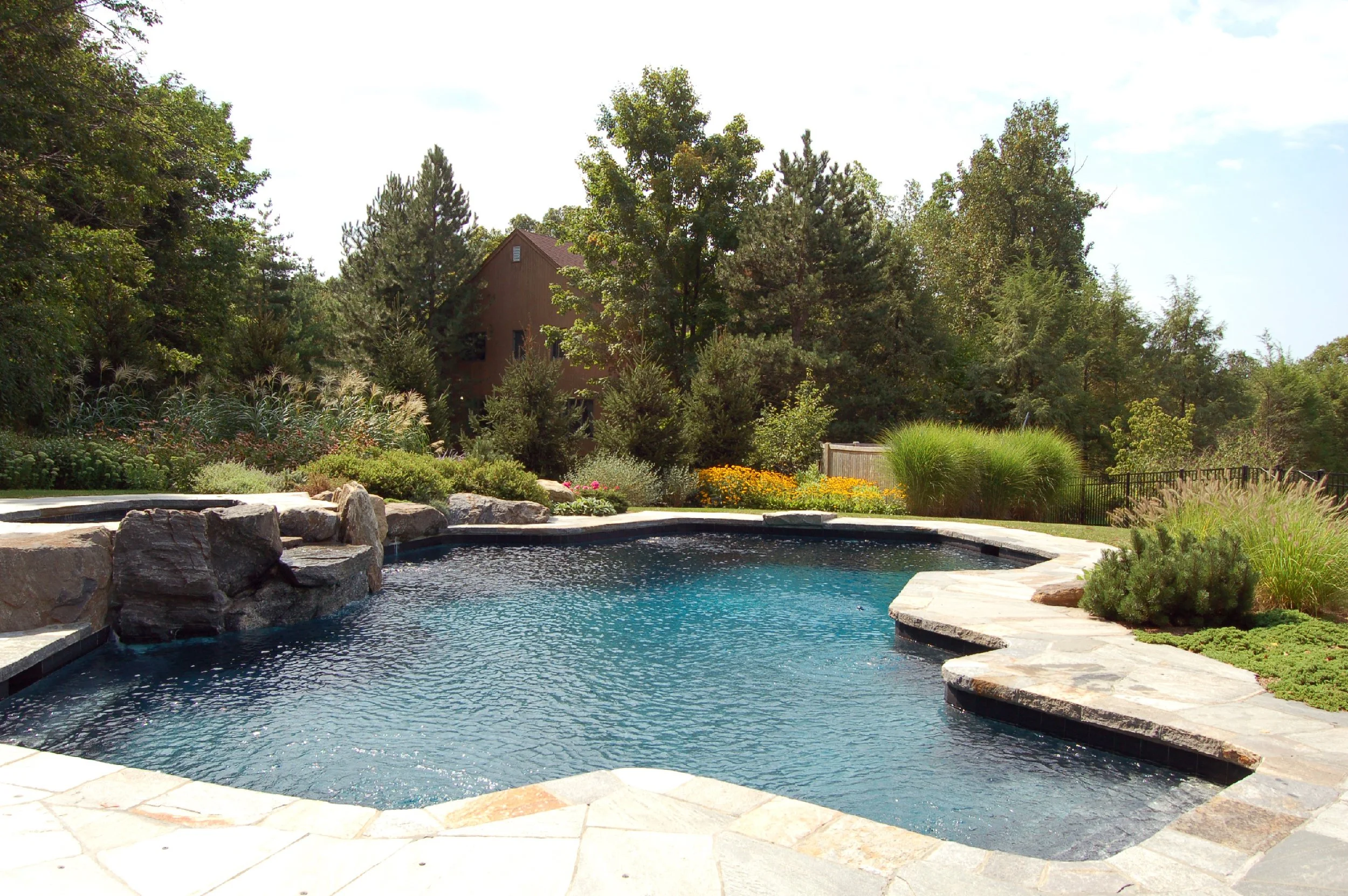 A backyard with a swimming pool surrounded by natural stone patio and lush green bushes, trees, and colorful flowers in the background.