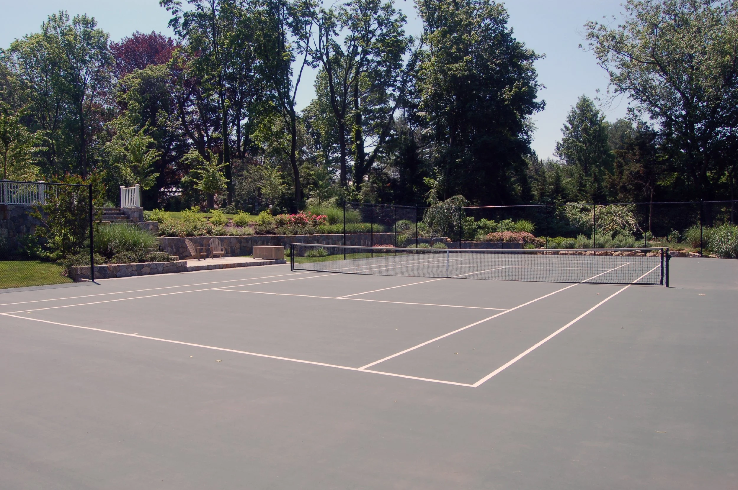 An empty tennis court with a net, surrounded by trees, bushes, and landscaped gardens in a park.