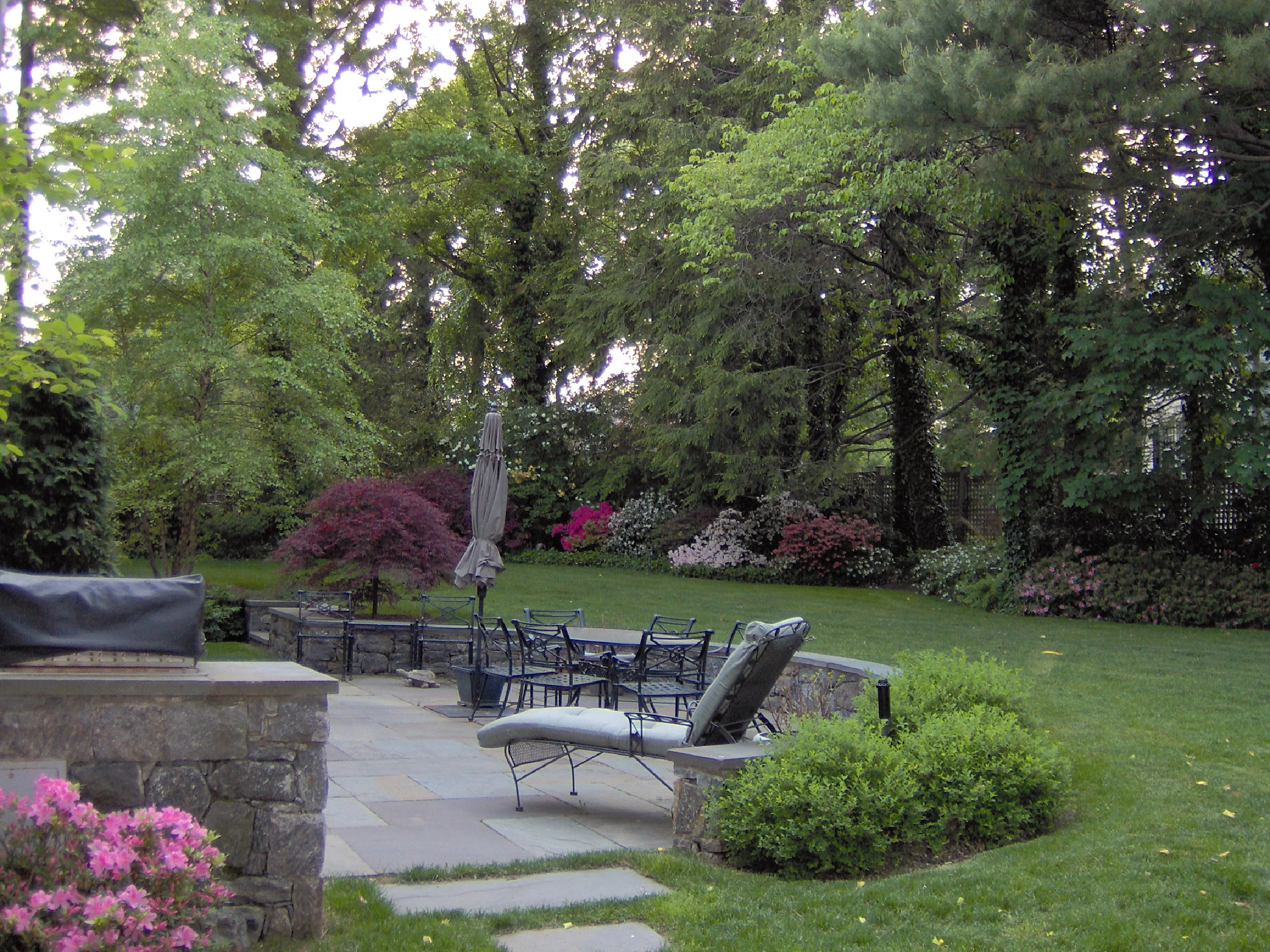 A backyard garden with trees, bushes, and flowering plants. There is a patio with tables, chairs, and lounge chairs, and an umbrella in the garden.