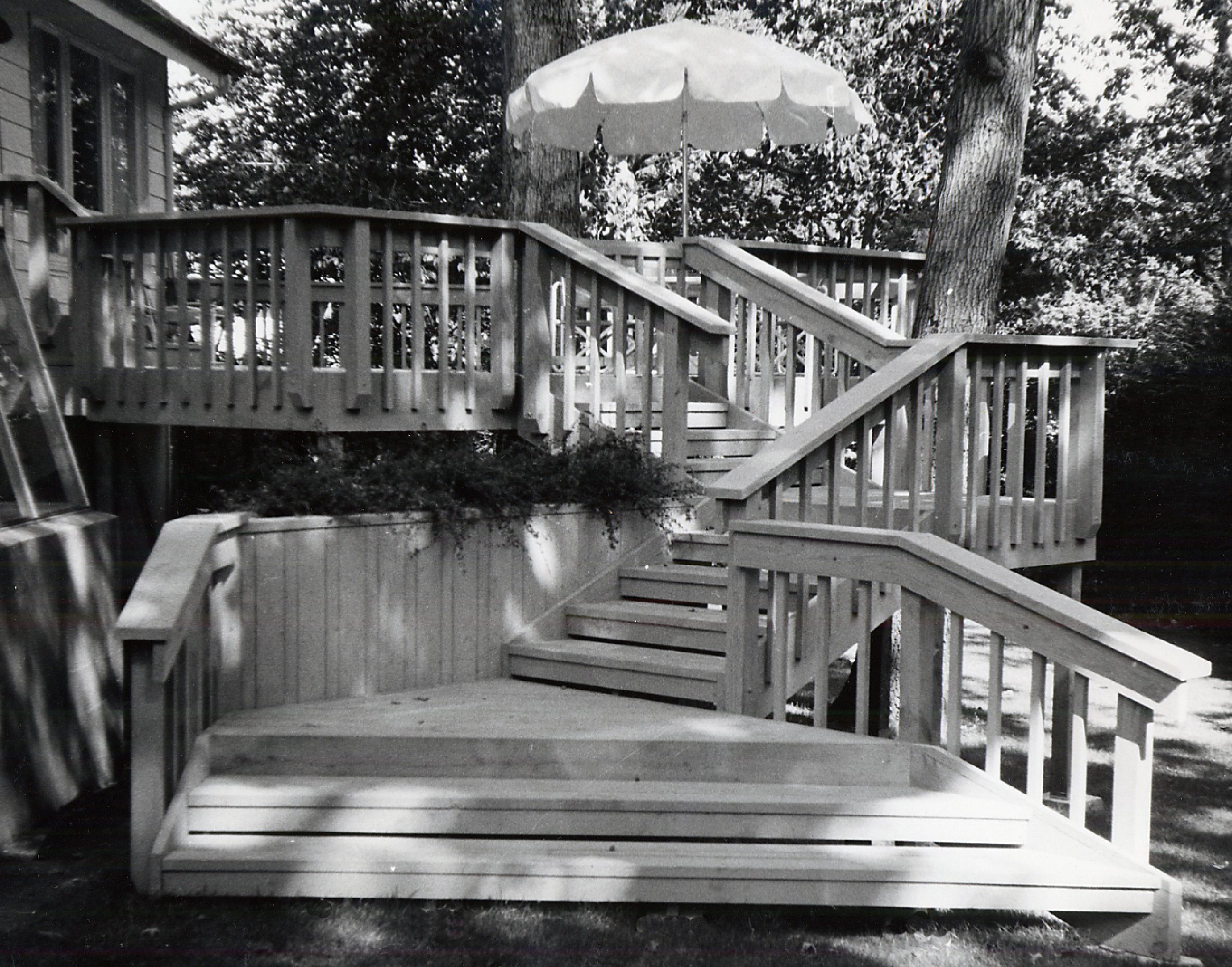 The image shows a multi-level wooden deck with stairs and railings, set outdoors with trees and foliage in the background. An umbrella is visible at the top of the deck.