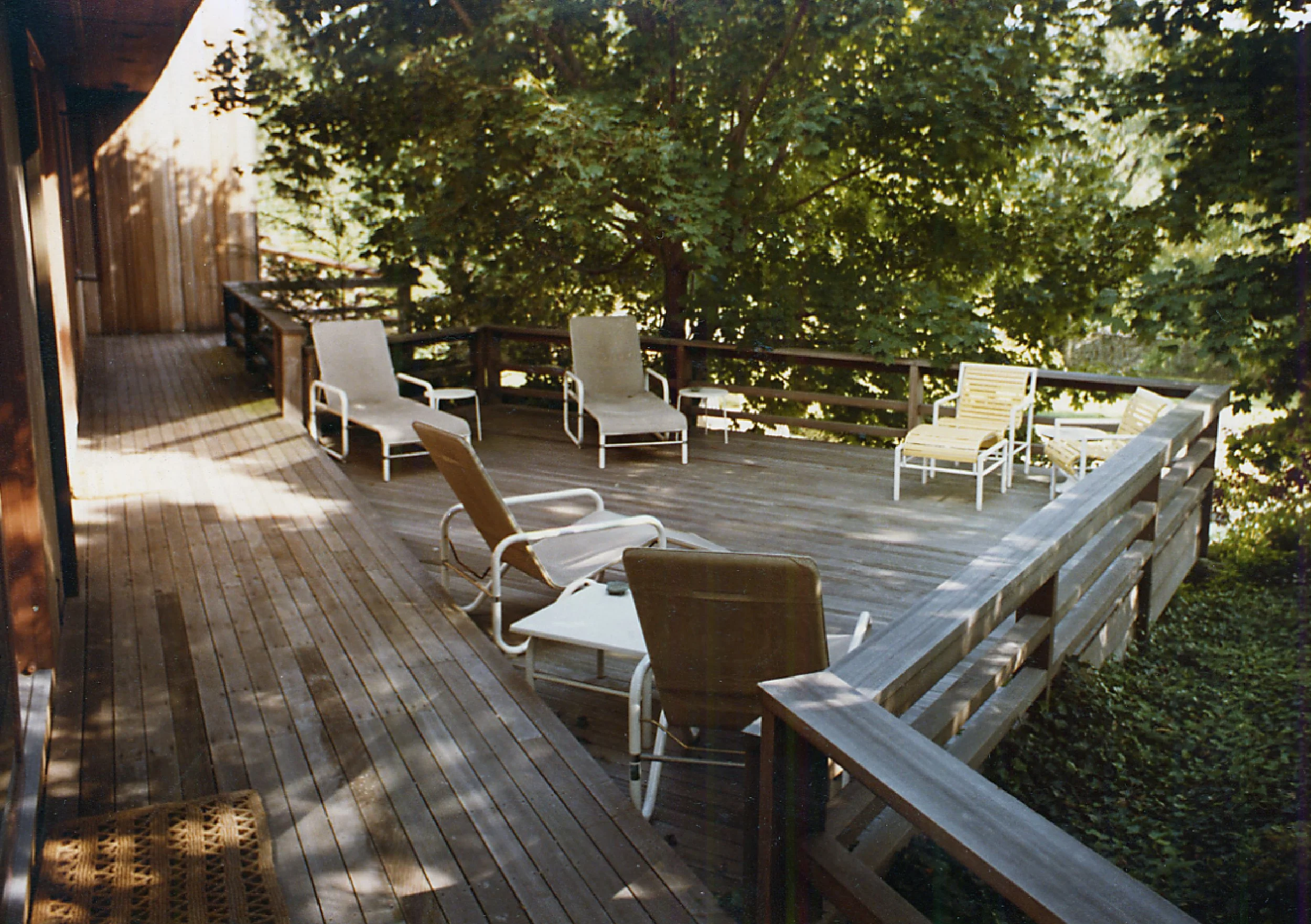 A wooden deck with several lounge chairs and small tables, surrounded by lush green trees.