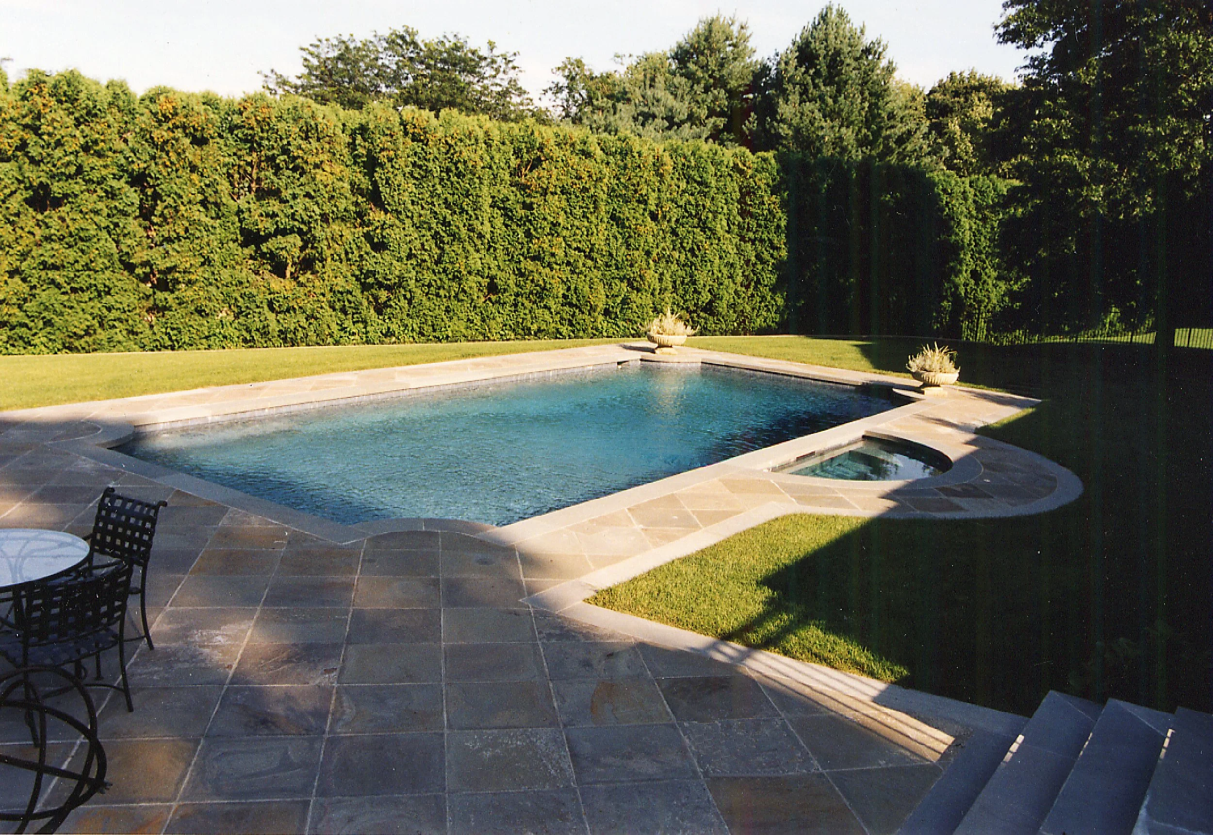 A backyard with a rectangular swimming pool, surrounded by a stone patio, with green grass and tall hedges and trees in the background.