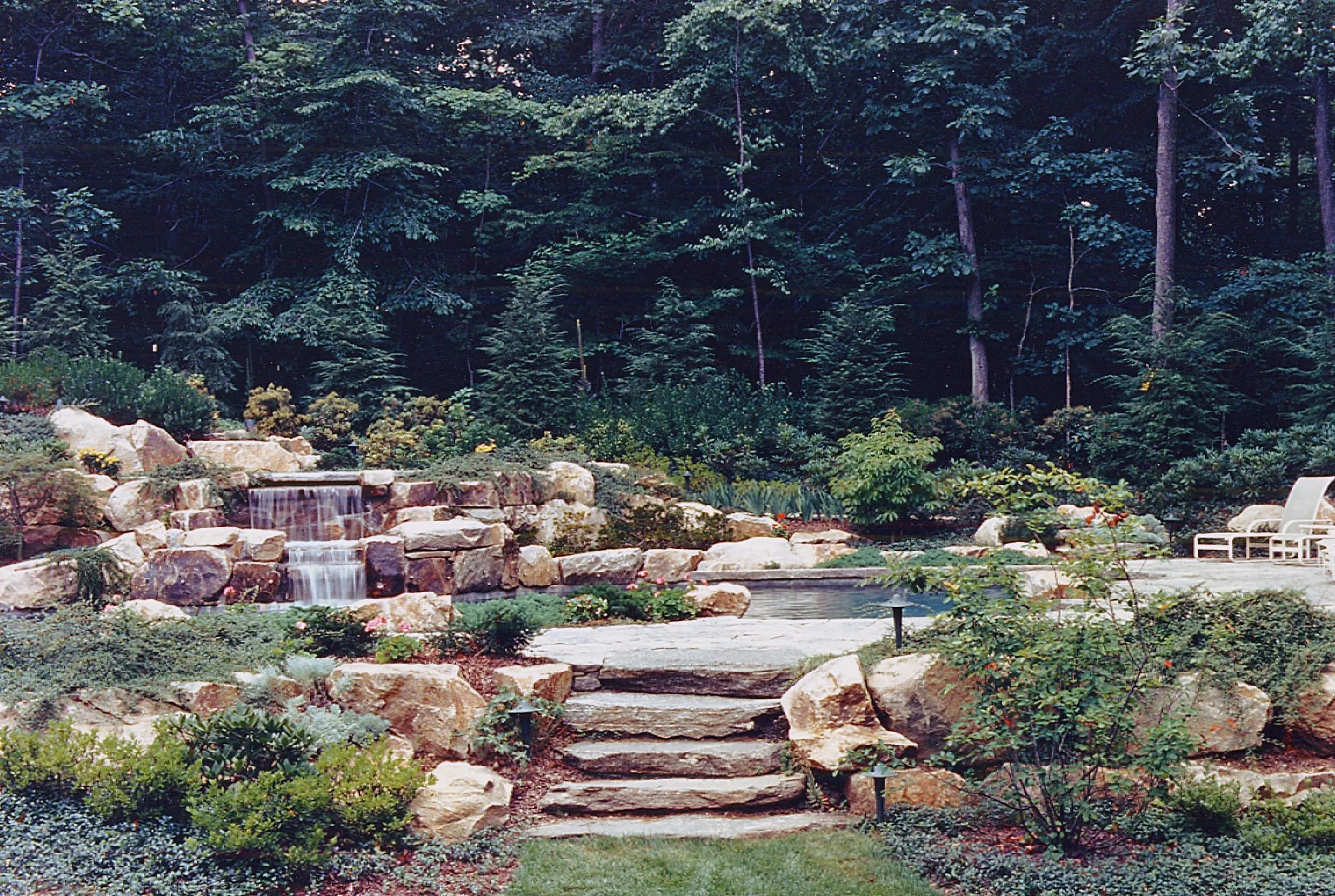 A landscaped backyard with a small waterfall flowing over rocks, stone steps leading to a patio, a garden with shrubs and flowers, and a wooded forest background.