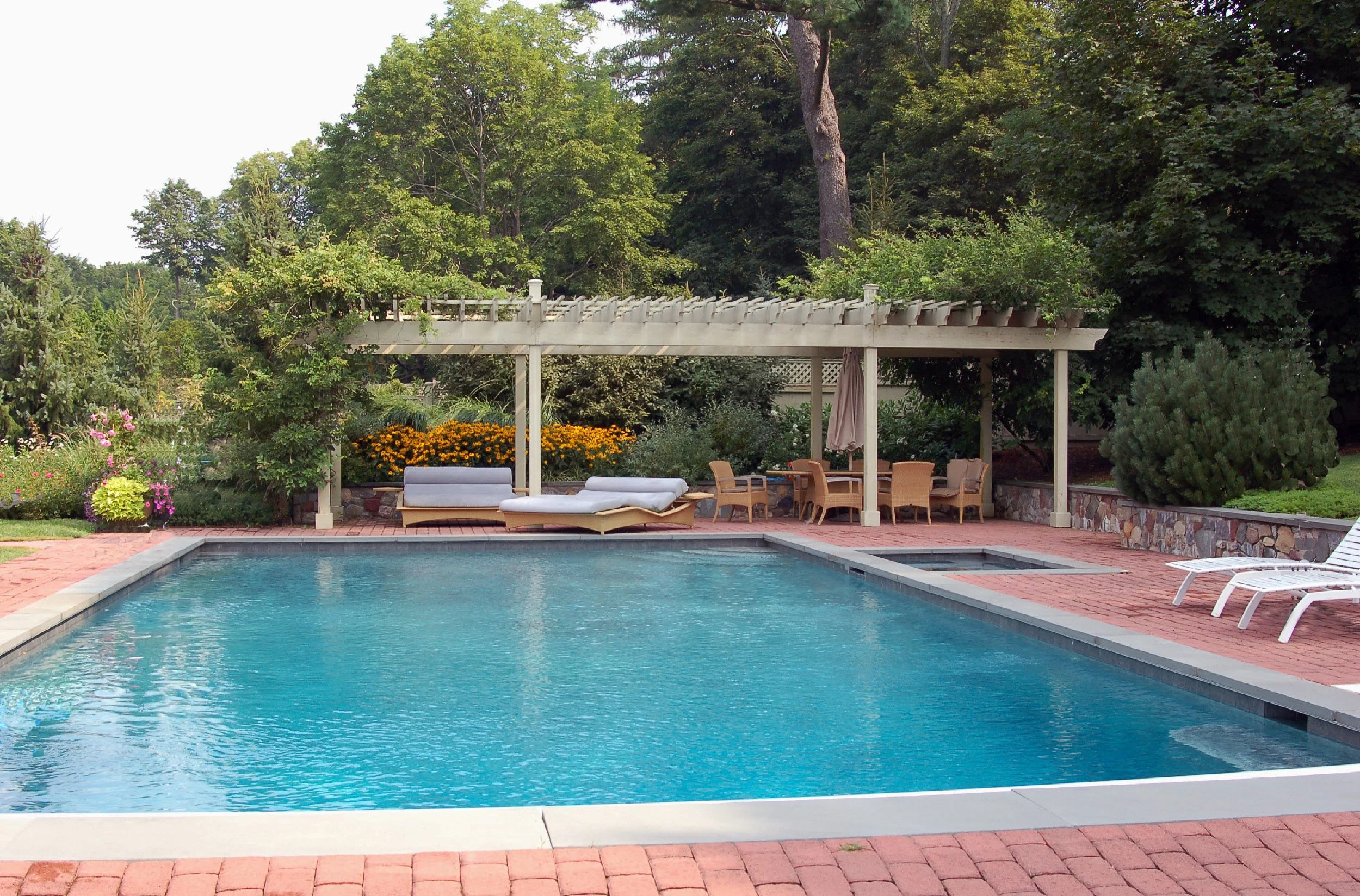 A backyard swimming pool with a white border, surrounded by a red brick patio. There is a shaded seating area with a pergola, outdoor furniture, lounge chairs, and lush greenery in the background.