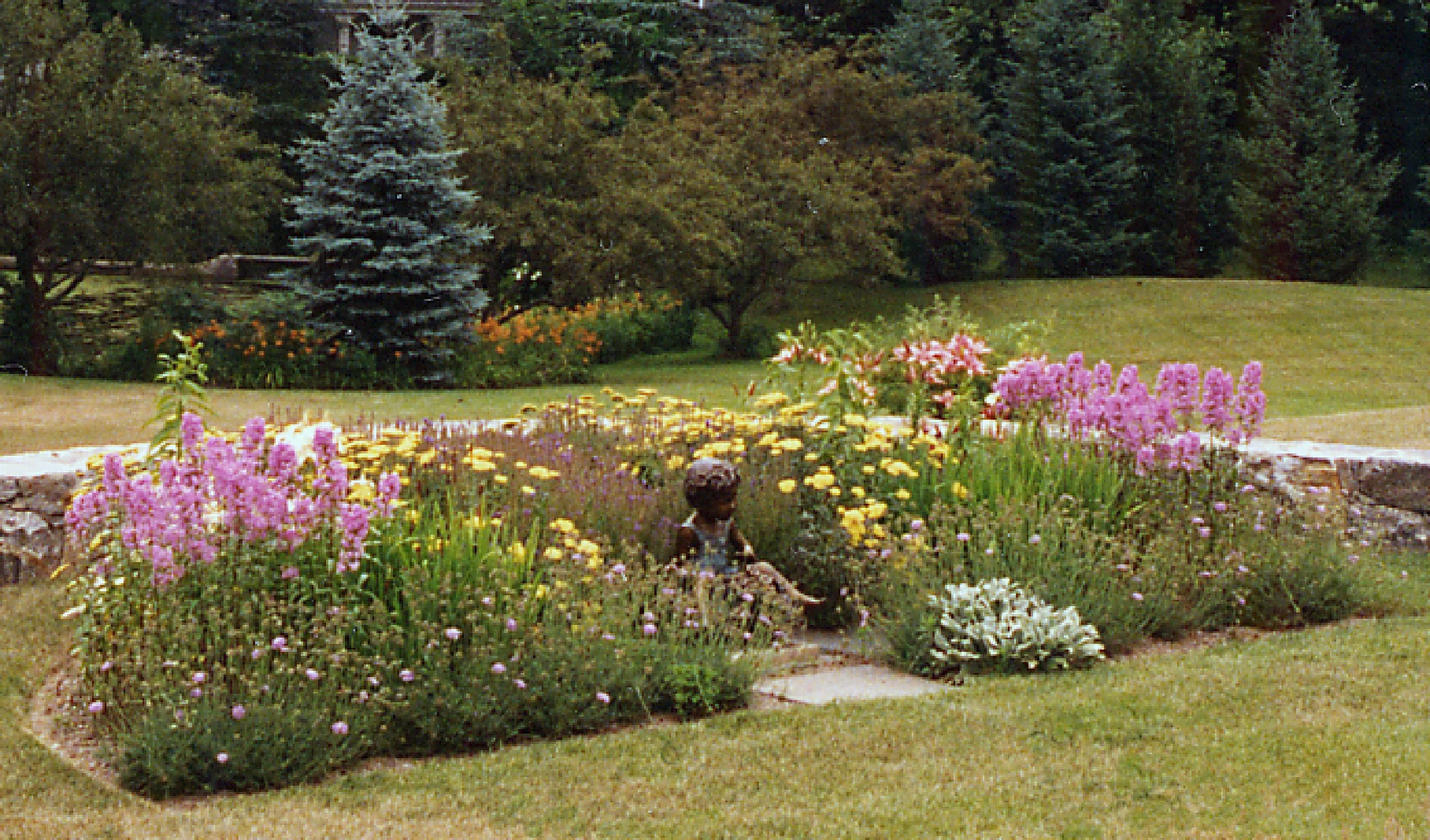 A landscaped garden with vibrant pink, yellow, and white flowers, a garden fountain with a statue in the center, and trees in the background.