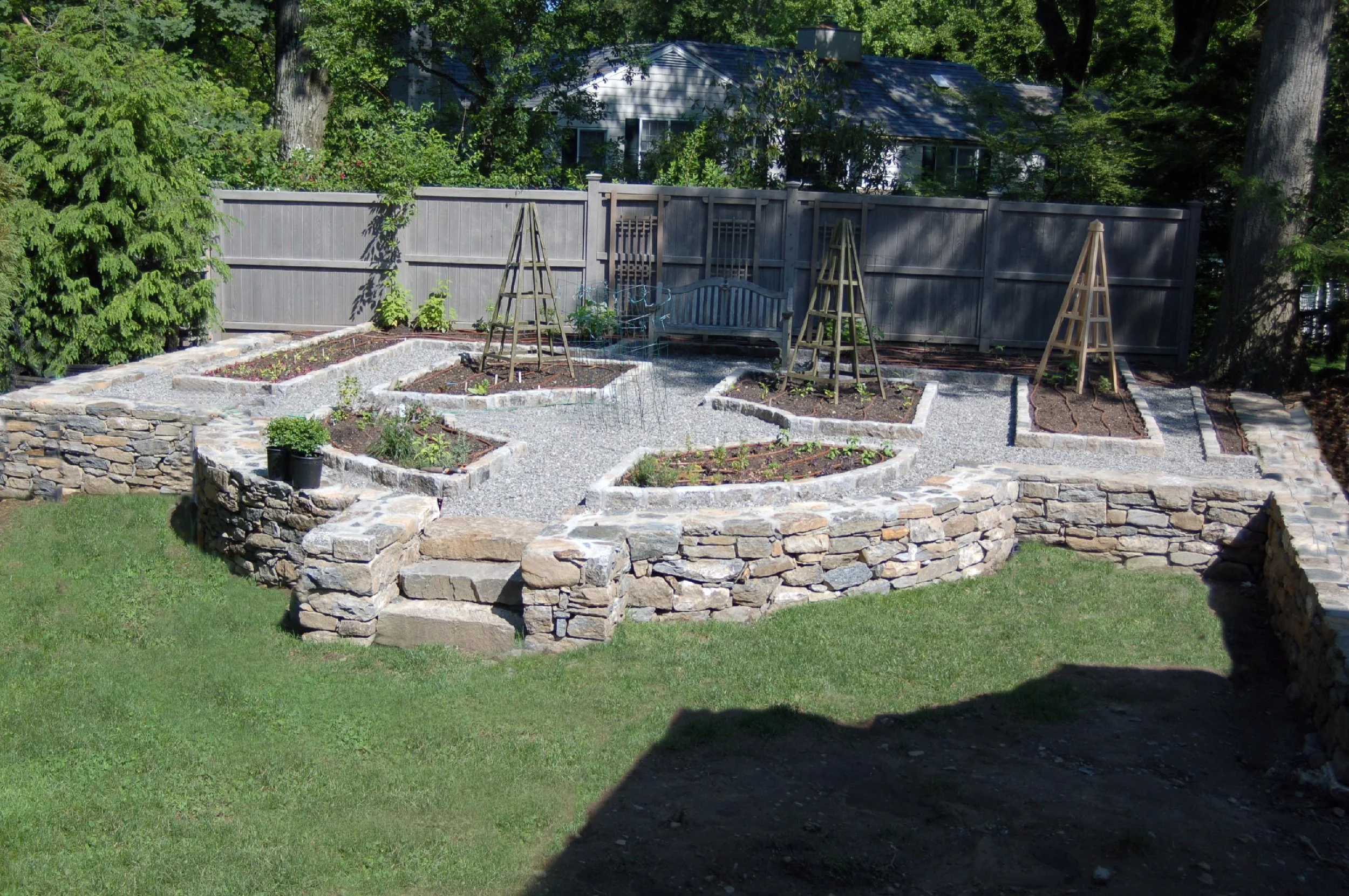 A landscaped backyard garden with raised garden beds, a stone retaining wall, a wooden fence, and trees in the background.