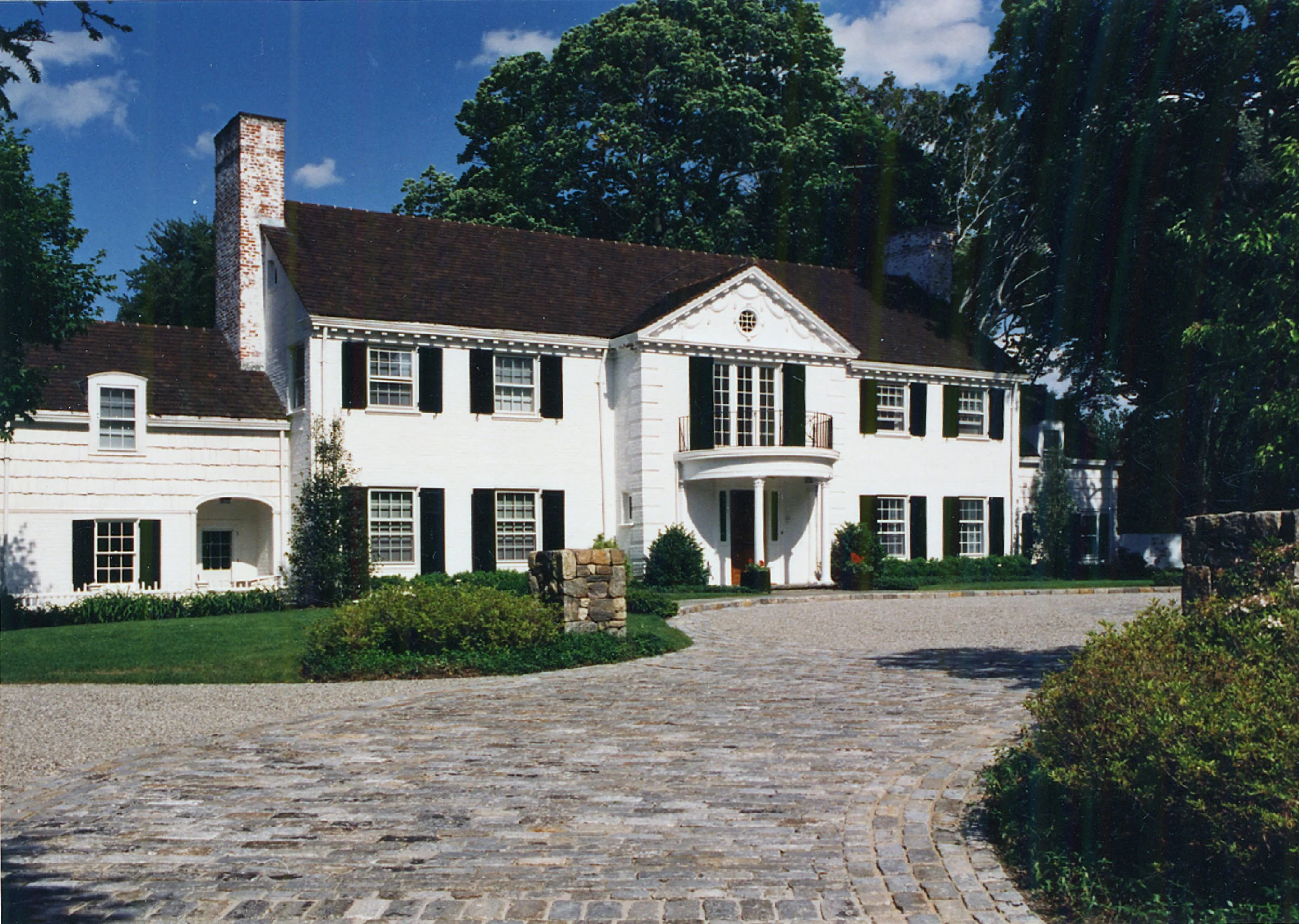 A large white two-story house with black shutters, a red-tiled roof, a chimney, and a small balcony above the front entrance. It is surrounded by a garden with bushes and a stone-paved driveway.