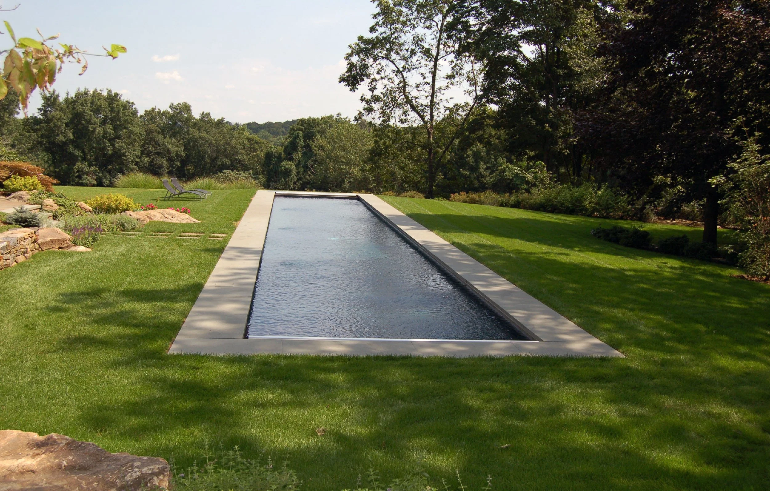 A backyard with a rectangular in-ground swimming pool surrounded by a concrete border, lush green lawn, trees, and garden plants, with a patio area and two lounge chairs in the background.