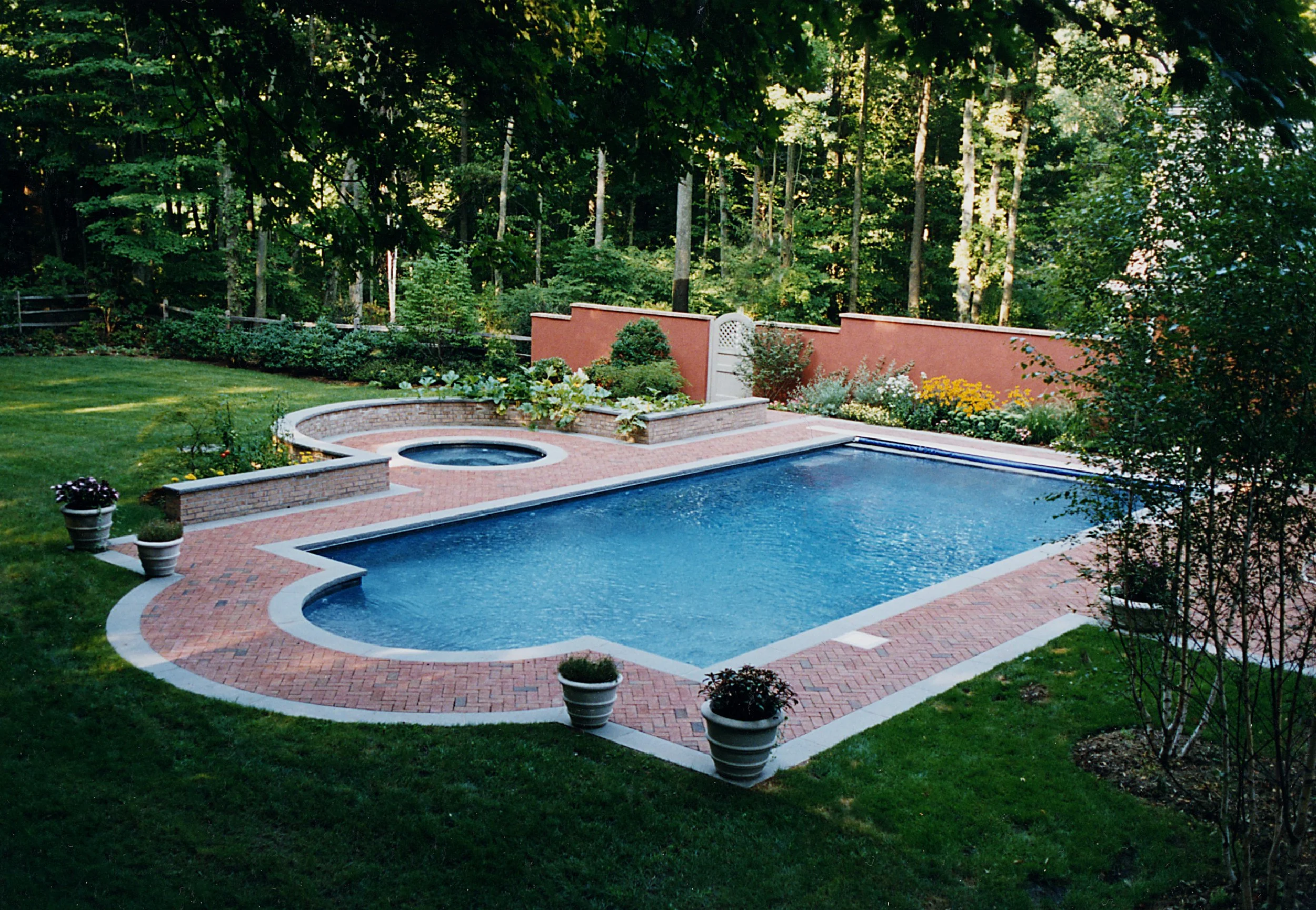 Residential backyard with a swimming pool surrounded by a brick patio, potted plants, and a landscaped garden with trees and flowers, enclosed by a red wall and natural woods.