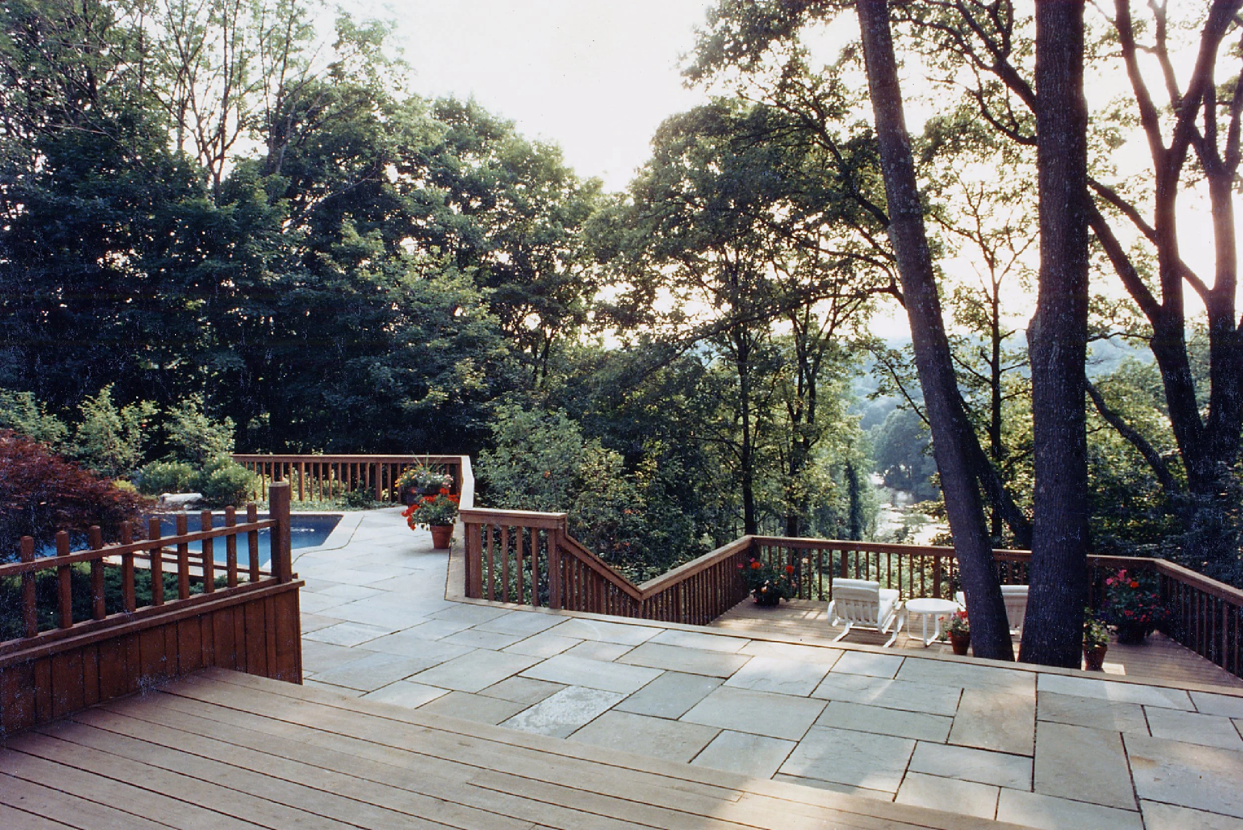 A backyard deck with stone and wood flooring, surrounded by wooden railings, with potted plants, garden seating, and trees with a view of a pond or lake in the distance.