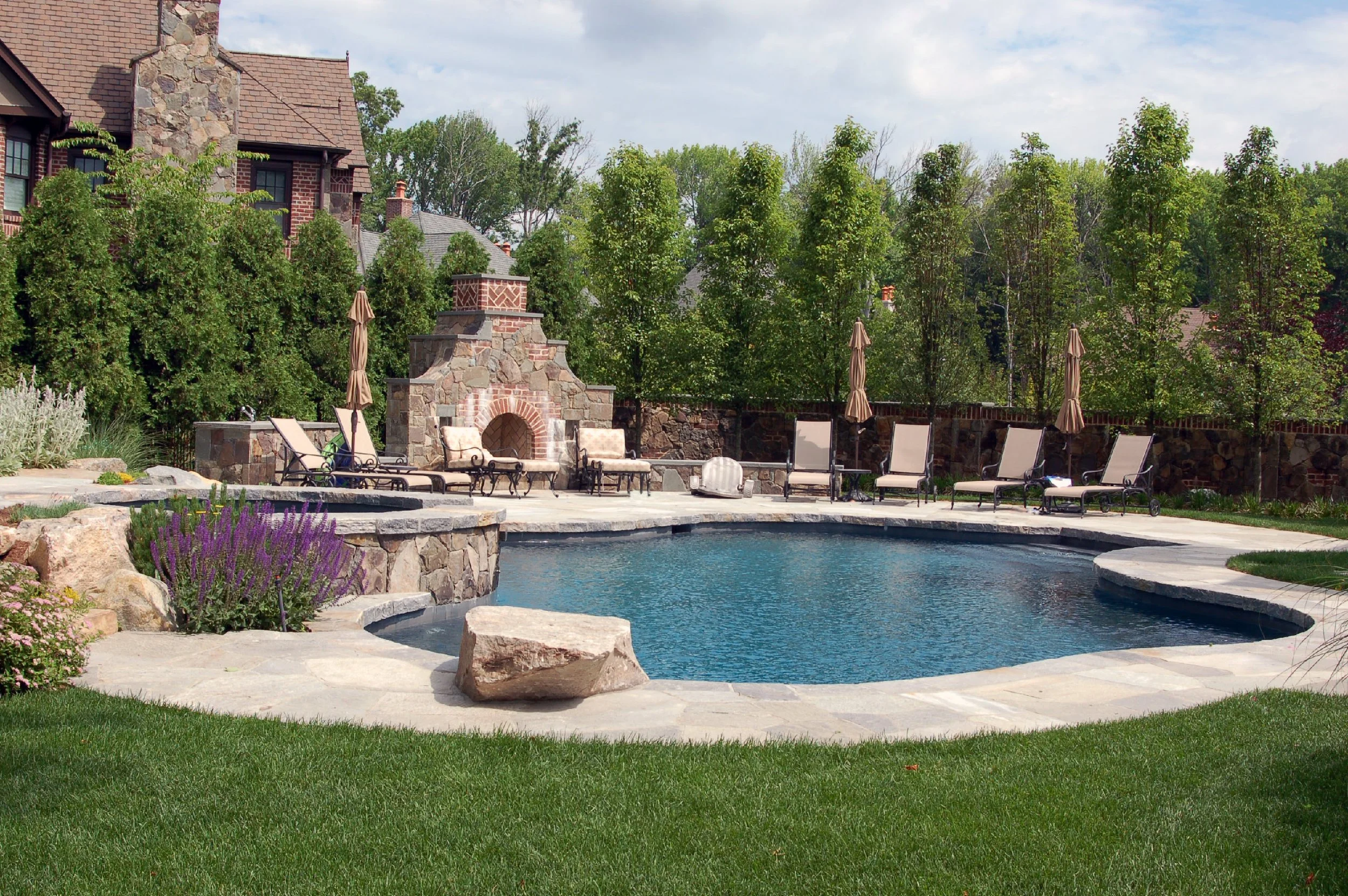 Backyard with a kidney-shaped swimming pool, surrounded by patio furniture, umbrellas, and a stone fireplace, with lush green trees and a house in the background.