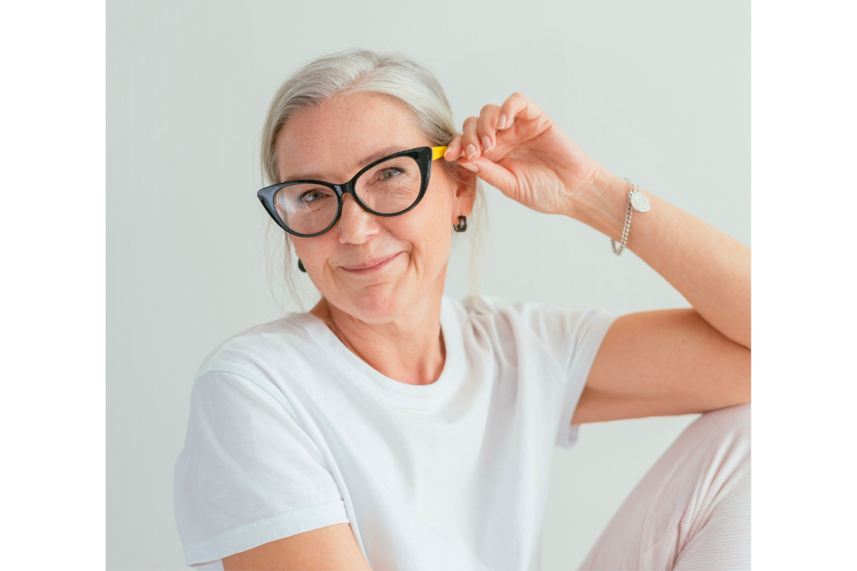 A smiling elderly woman with gray hair, wearing black glasses, a white t-shirt, and jewelry, holding her glasses with one hand.