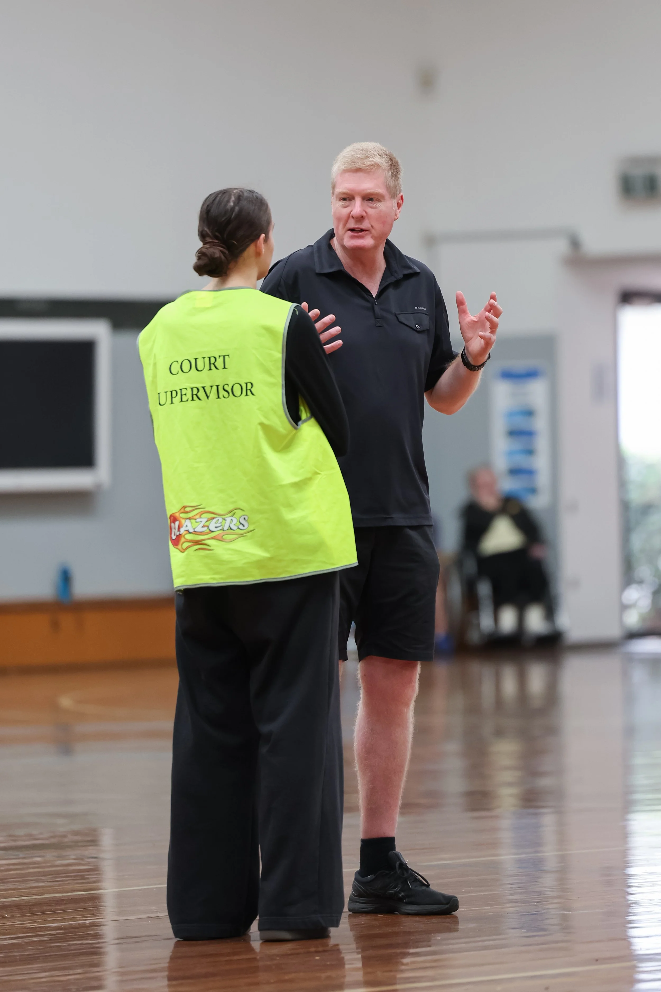 A woman wearing a yellow vest labeled 'Court Supervisor' is talking to a man in athletic shorts and black shoes in a gymnasium.