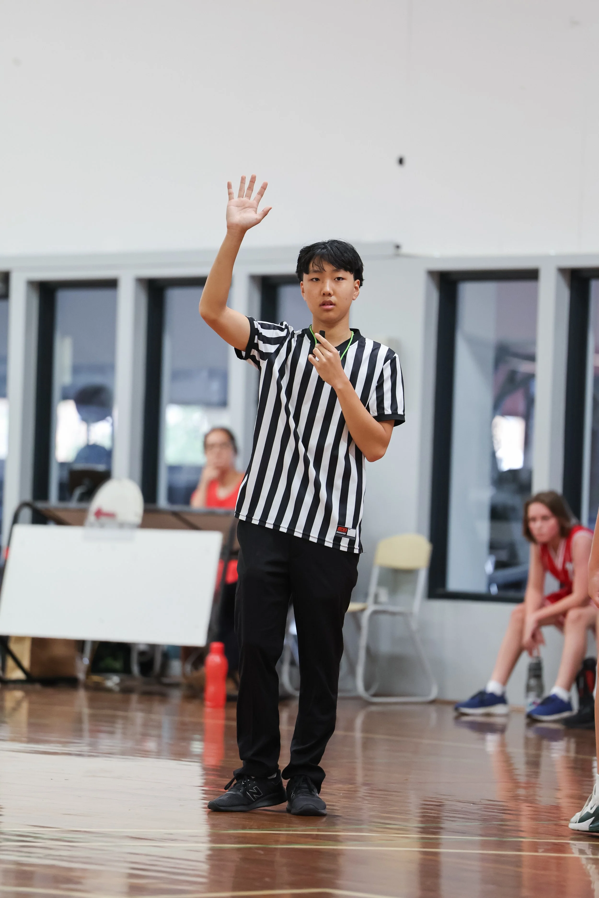 A young male referee in black and white striped shirt standing on a gym floor, raising his right hand, with people seated on chairs behind him.