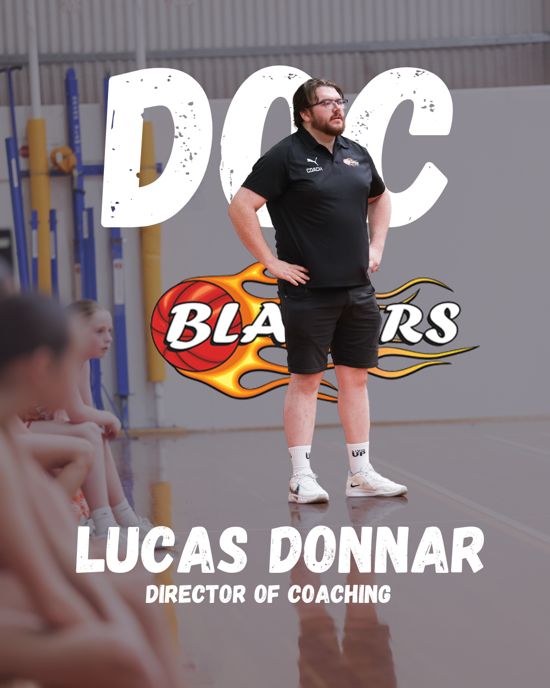 A basketball coach standing on a gym floor with children sitting nearby, in front of a wall with a large 'DOC' and 'BLAZERS' logo.