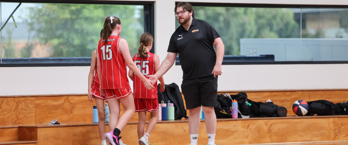 A coach shaking hands with two young female basketball players in red and white uniforms, inside a gymnasium.