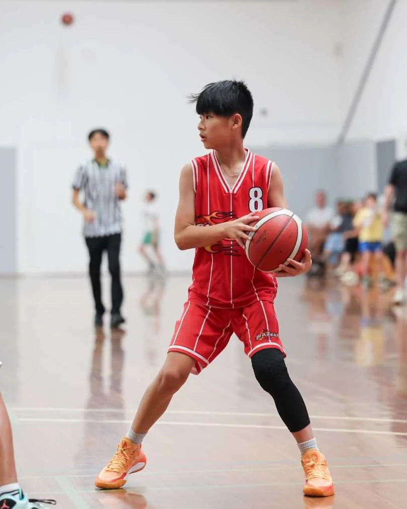 A young boy in a red basketball uniform holding a basketball on an indoor court, with spectators and a referee in the background.