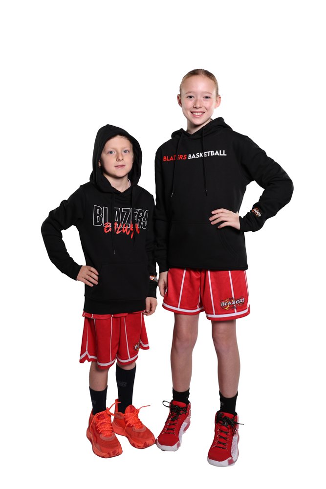 Two young girls dressed in Portland Trail Blazers basketball gear, standing together against a white background. The taller girl on the right wears a black hoodie and red basketball shorts, while the shorter girl on the left wears a similar hoodie and shorts. Both girls are wearing red sneakers.