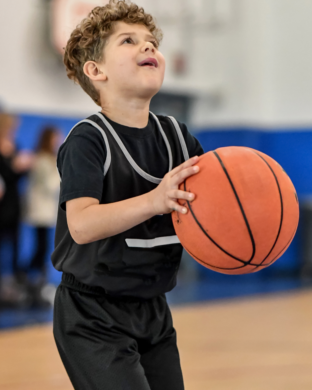 Young boy in a black sports uniform holding a basketball on an indoor gym court.