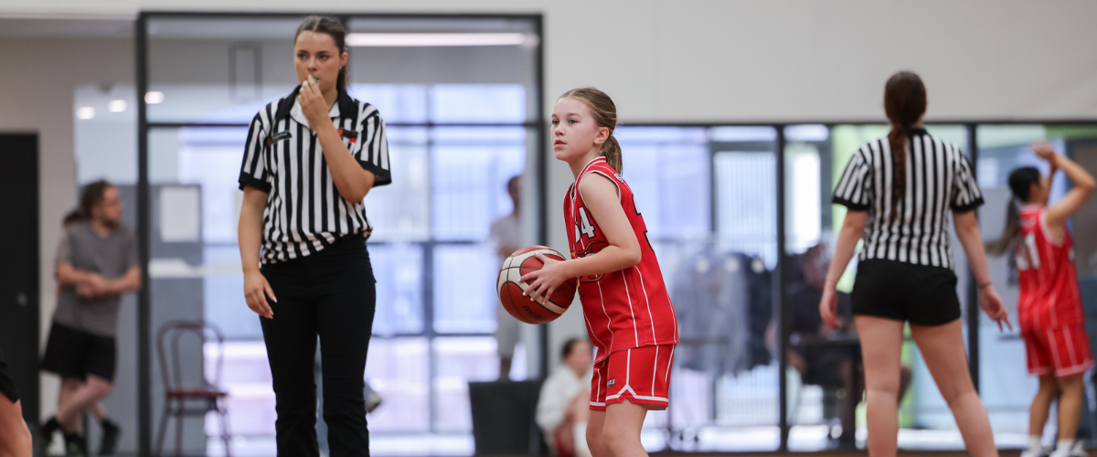 A young girl in a red basketball uniform holding a basketball on an indoor court during a youth basketball game, with referees and other players present.