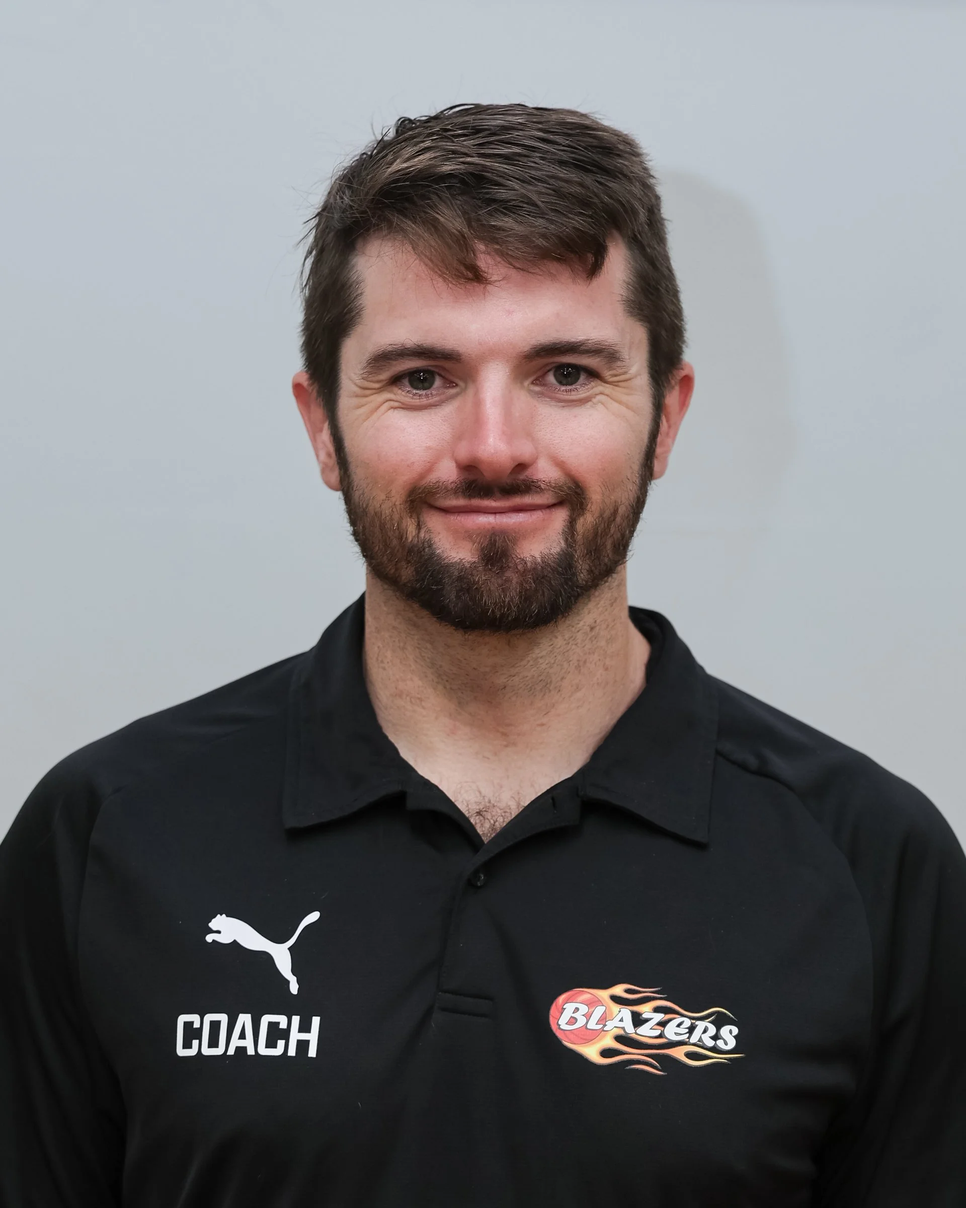 A man with brown hair and a beard wearing a black polo shirt with the Puma logo, the word 'COACH,' and a logo with a flaming basketball and the word 'BLAZERS.'