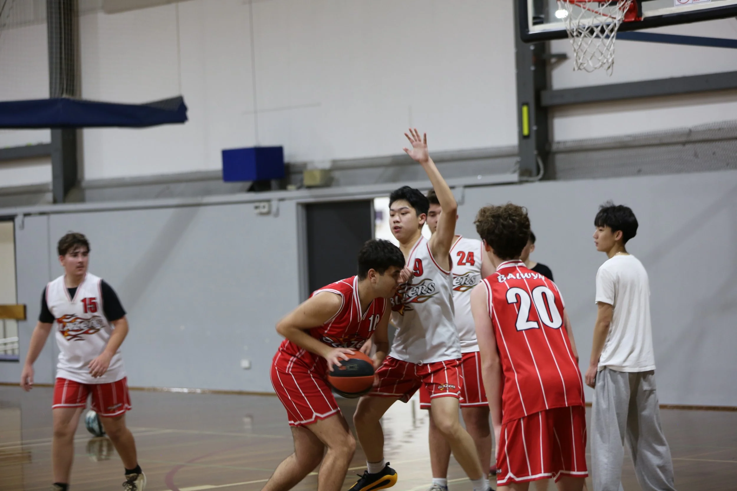 Young male basketball players in red and white uniforms playing on an indoor court, some gathered near the basketball hoop, with a few standing and others crouching.
