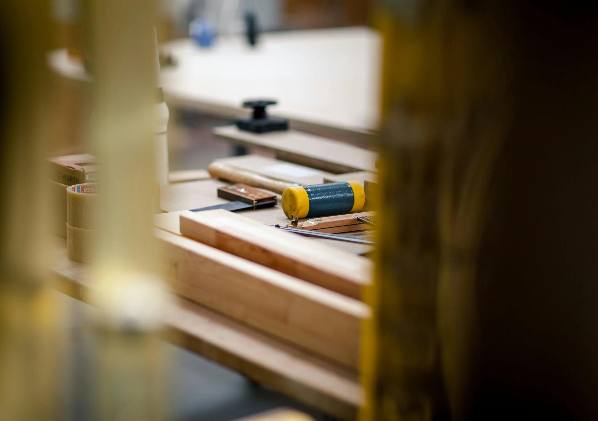 Woodworking workshop tools and materials on a workbench, partially obscured by blurred yellow structures in the foreground.